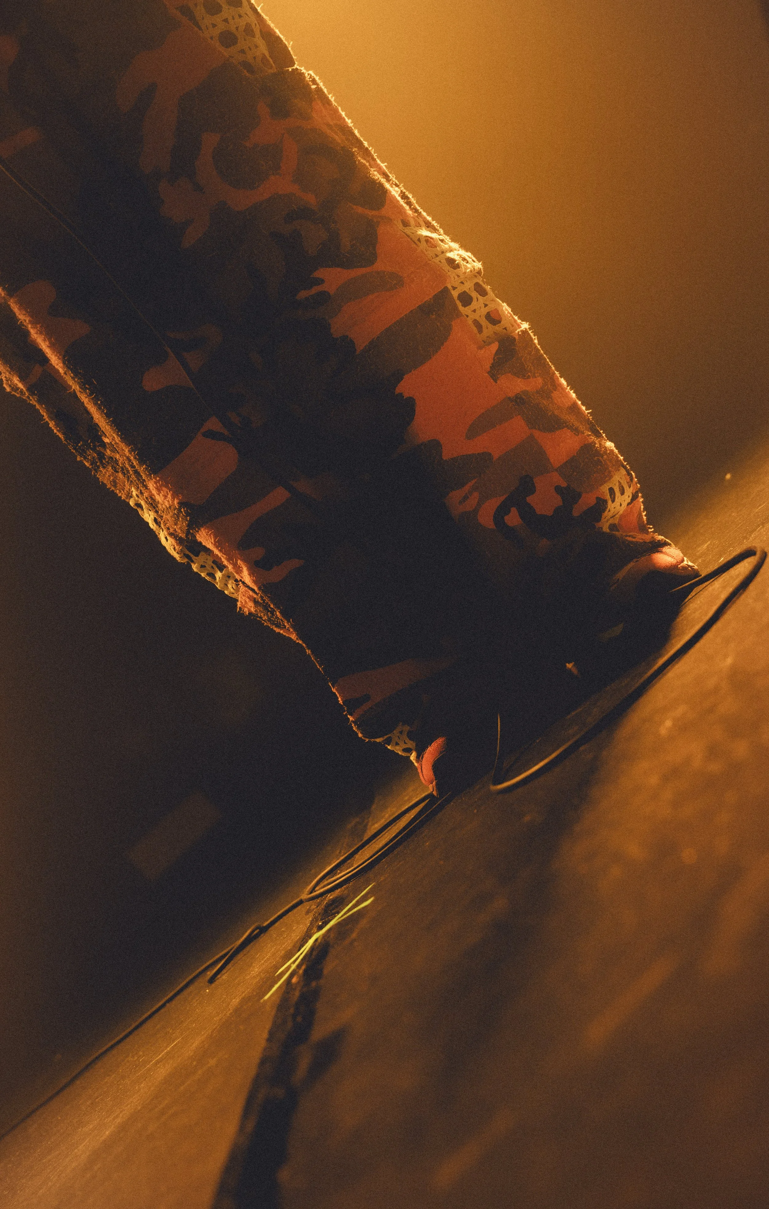 Close-up of a sneaker with orange and black camouflage pattern, illuminated by warm orange lighting, on a dark floor with visible cords or wires.