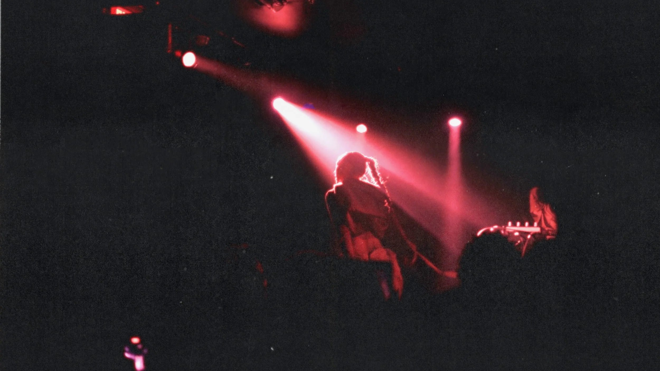 A person performing on stage under red lighting, with a guitarist nearby, and a dark background.