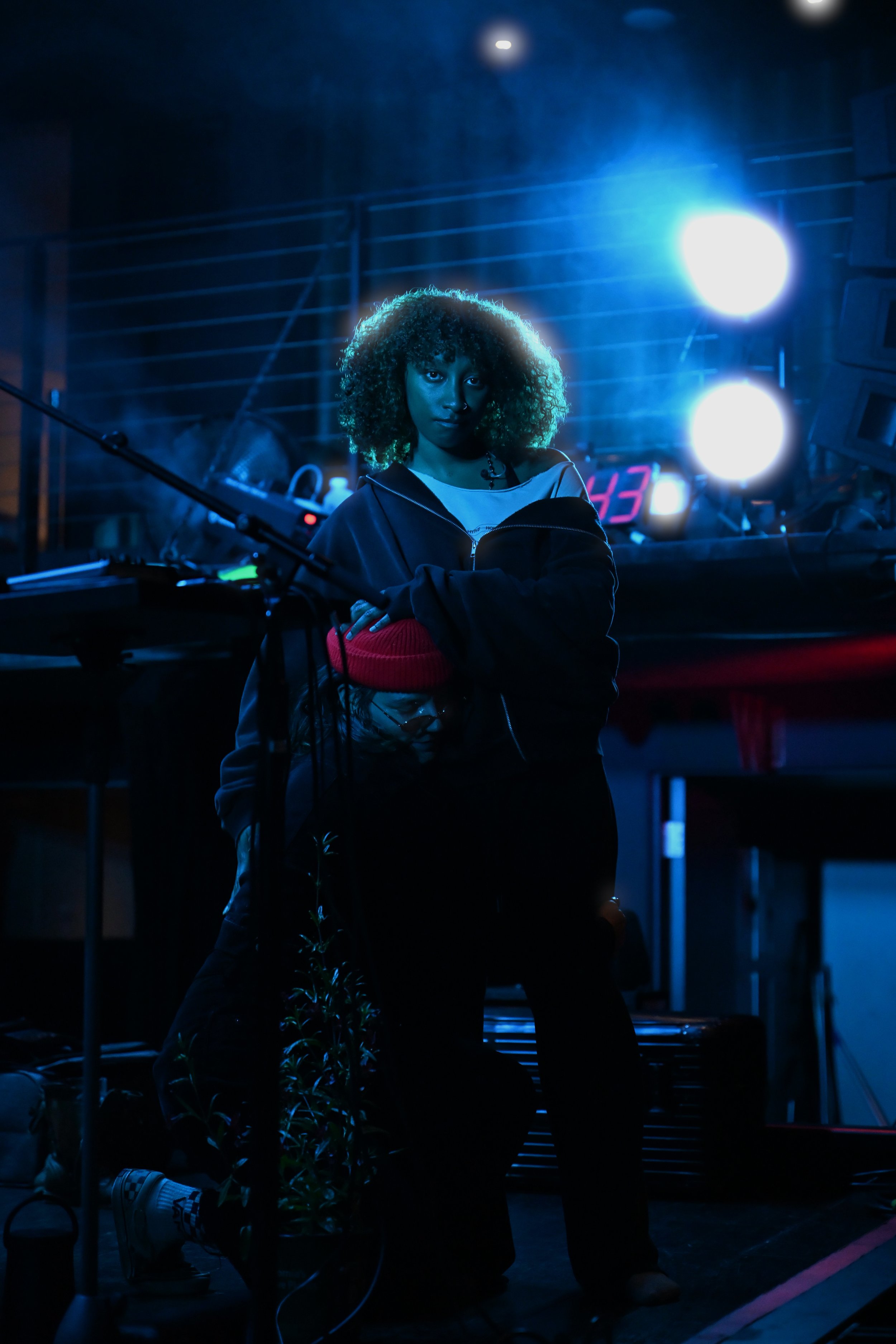 A woman with curly hair standing in a dimly lit room with blue and white lights, looking directly at the camera.