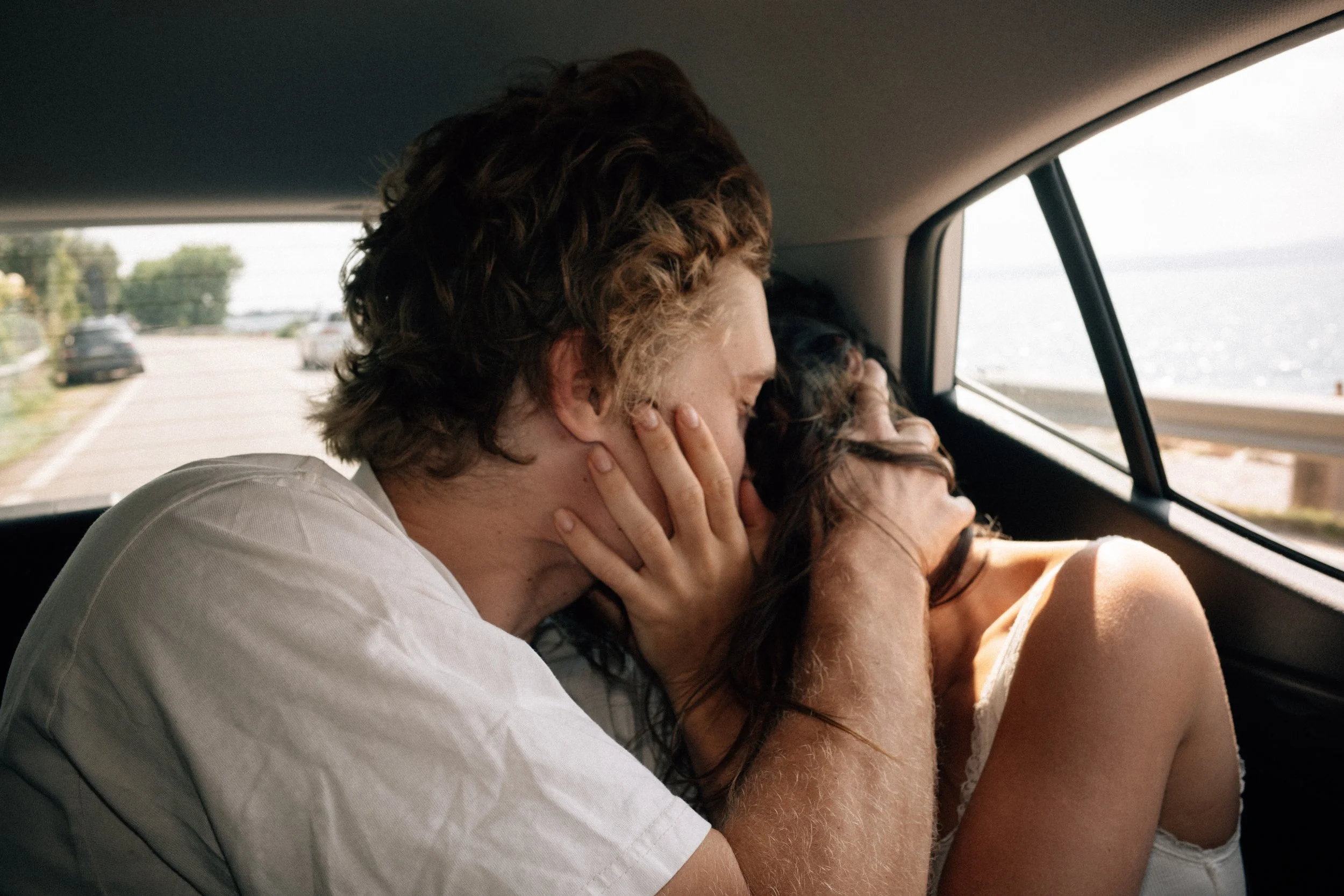 A couple is sharing an intimate kiss inside a car while parked by the water, with the man holding the woman gently.