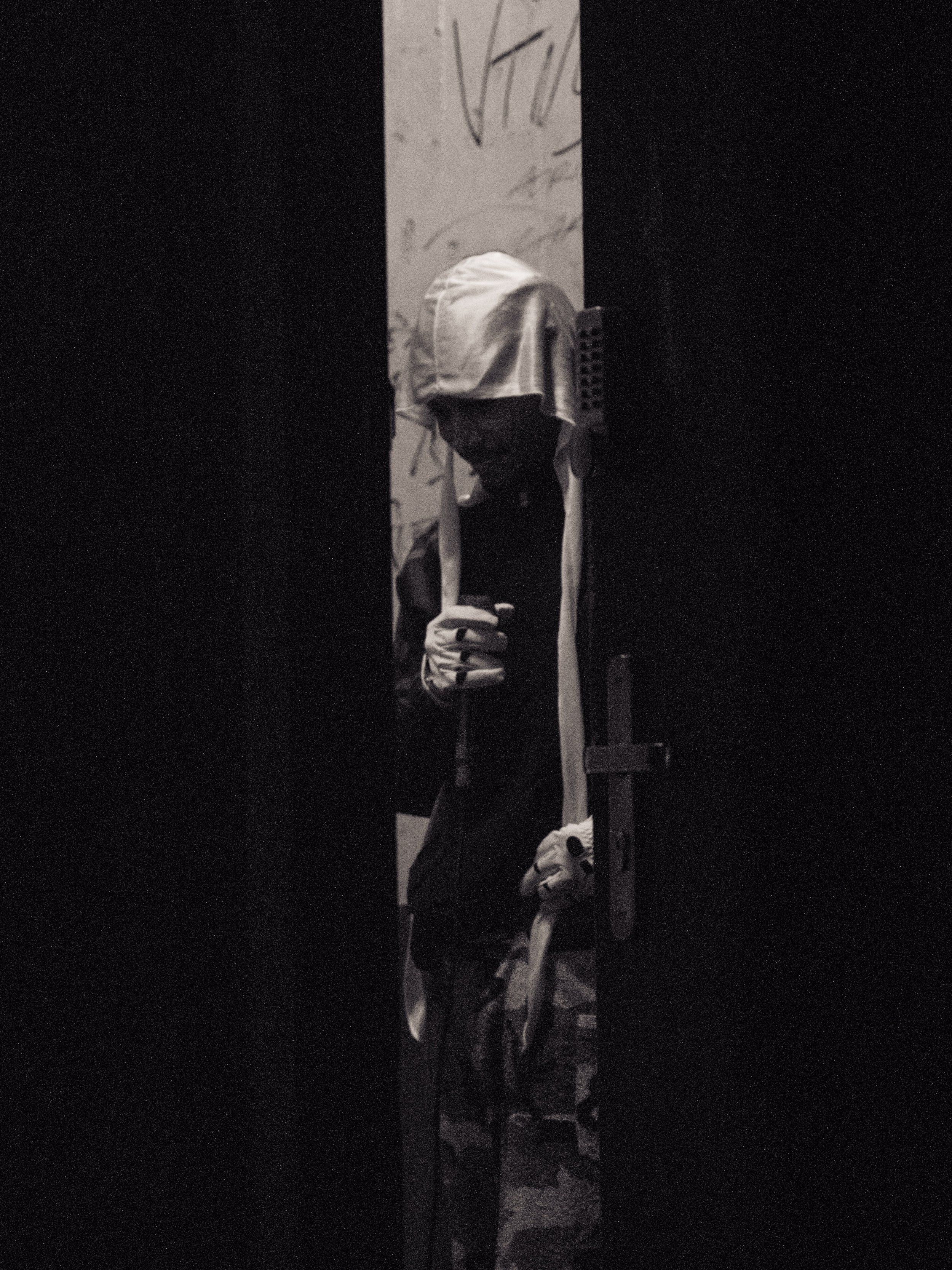 A person wearing a white face covering and gloves holding a piece of paper or a sign, partially hidden behind a dark piece of furniture or structure, with a background of a wall with graffiti or markings.