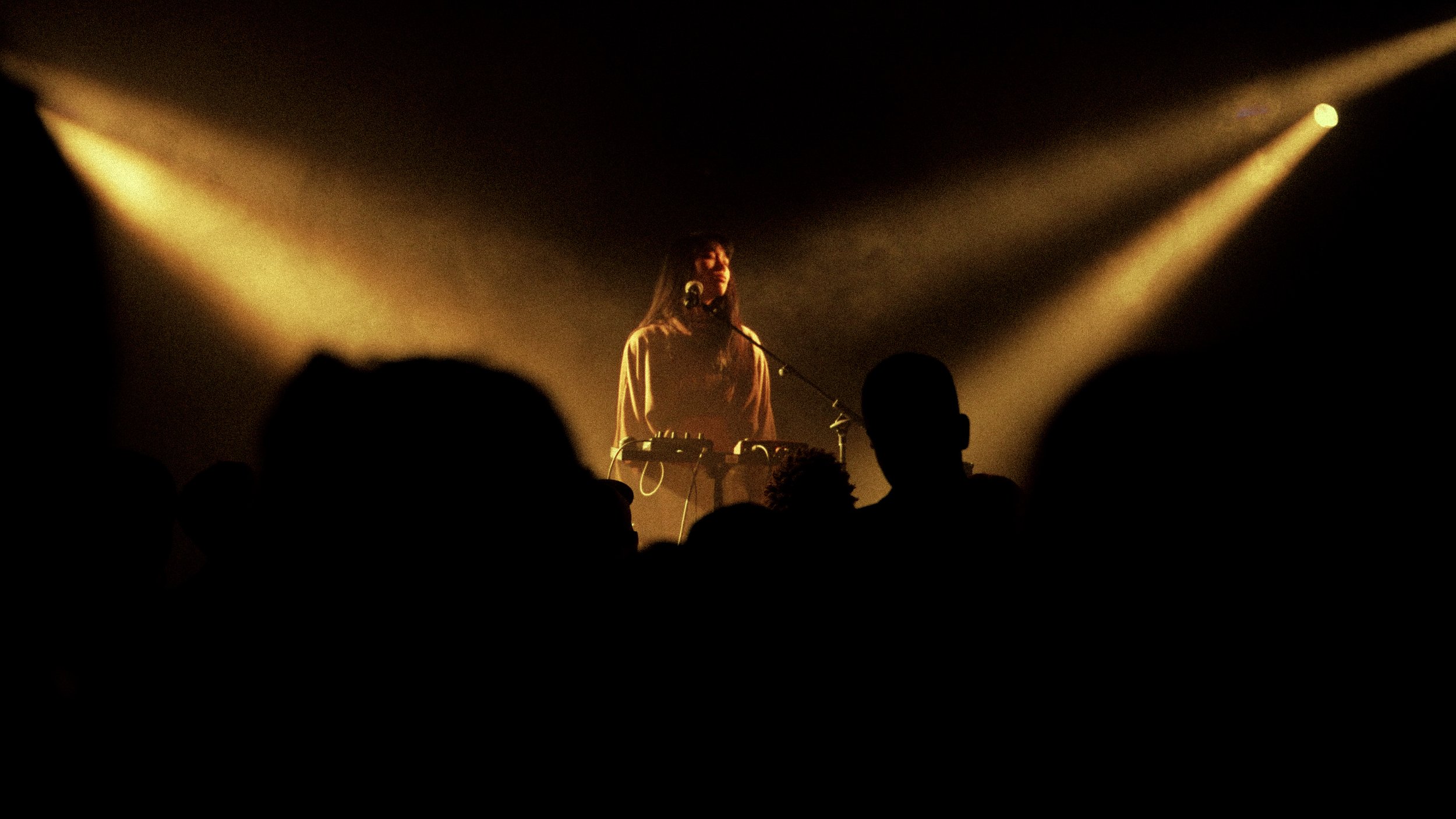 A person performing on stage with a music setup, illuminated by two bright stage lights, in front of a dark background, with audience silhouettes visible in the foreground.