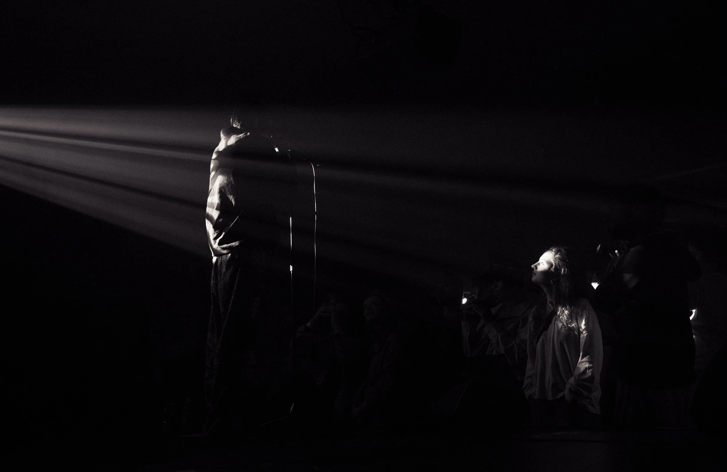 A person standing with head bowed, surrounded by beams of light in a dark environment, facing a seated woman with long hair, illuminated by light.