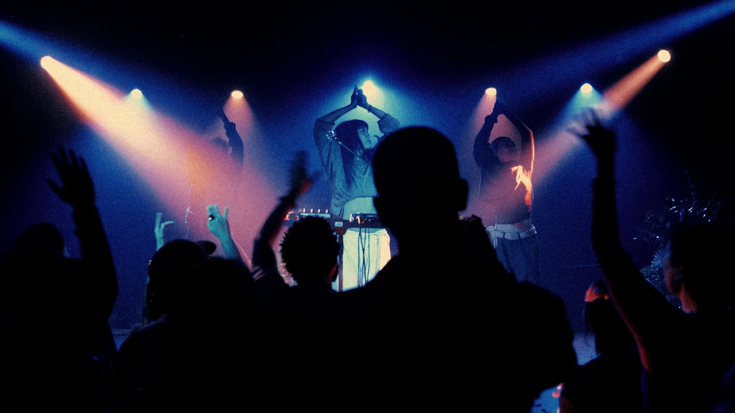 Silhouettes of people dancing and raising their hands at a concert or party with colorful stage lights.