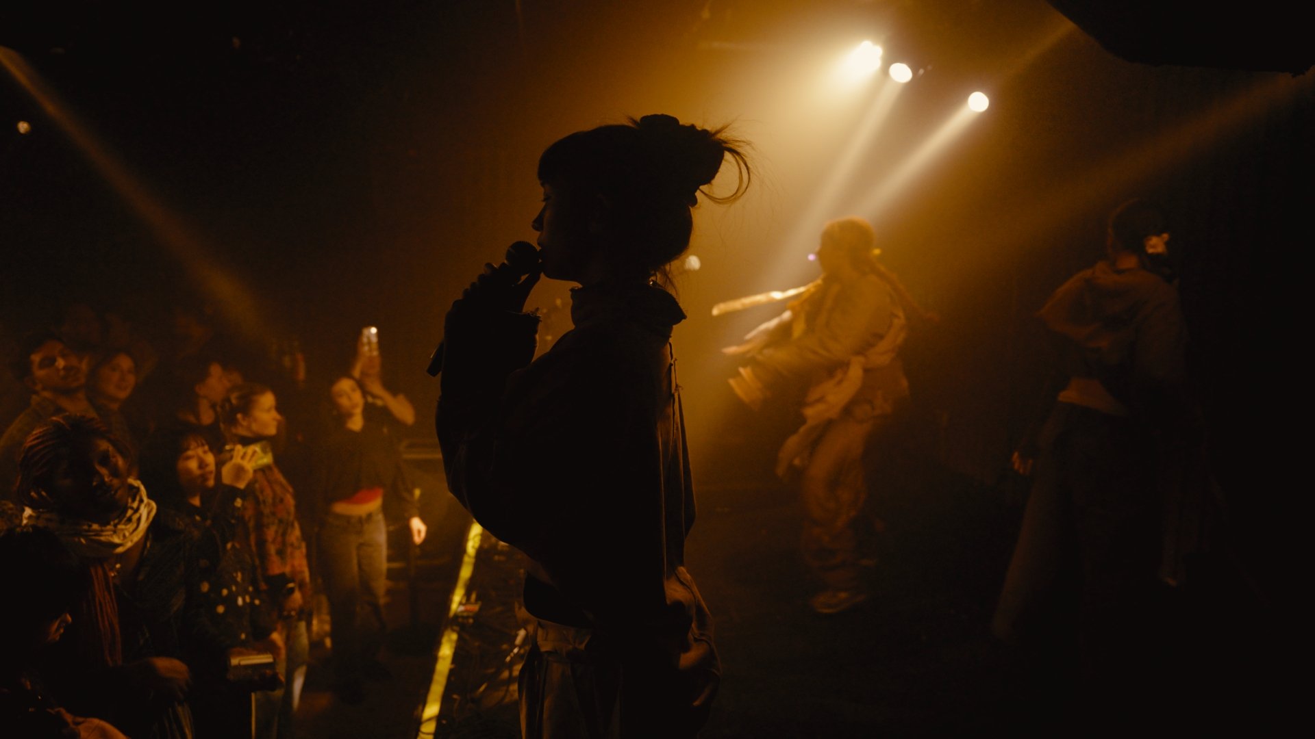 Silhouette of a person holding a microphone on stage, with a dance performance in the background, and an audience watching in a dimly lit setting with stage lights.