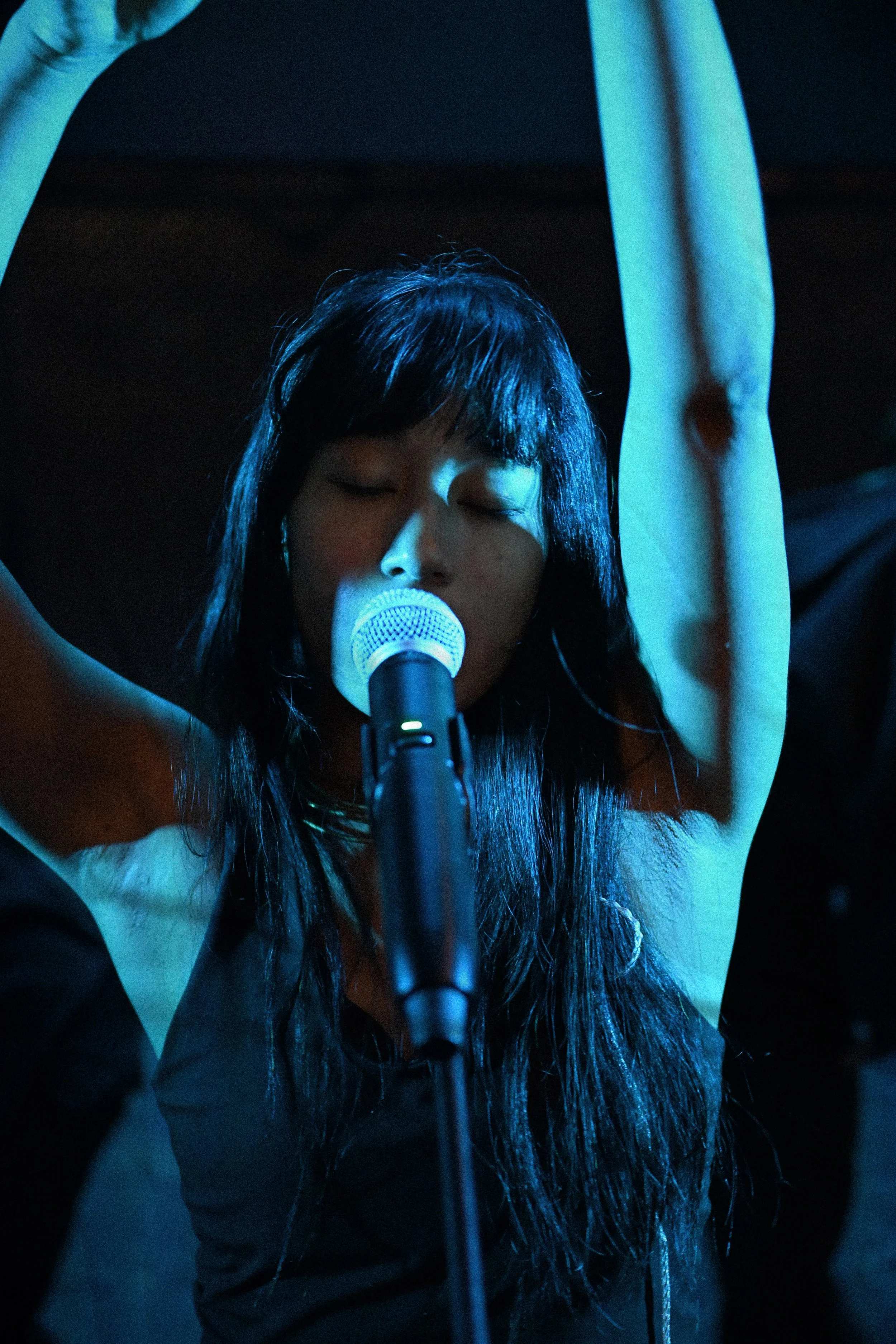 A person singing into a microphone with eyes closed, arms raised, long dark hair, wearing a black and white outfit, in a dark setting with blue lighting.
