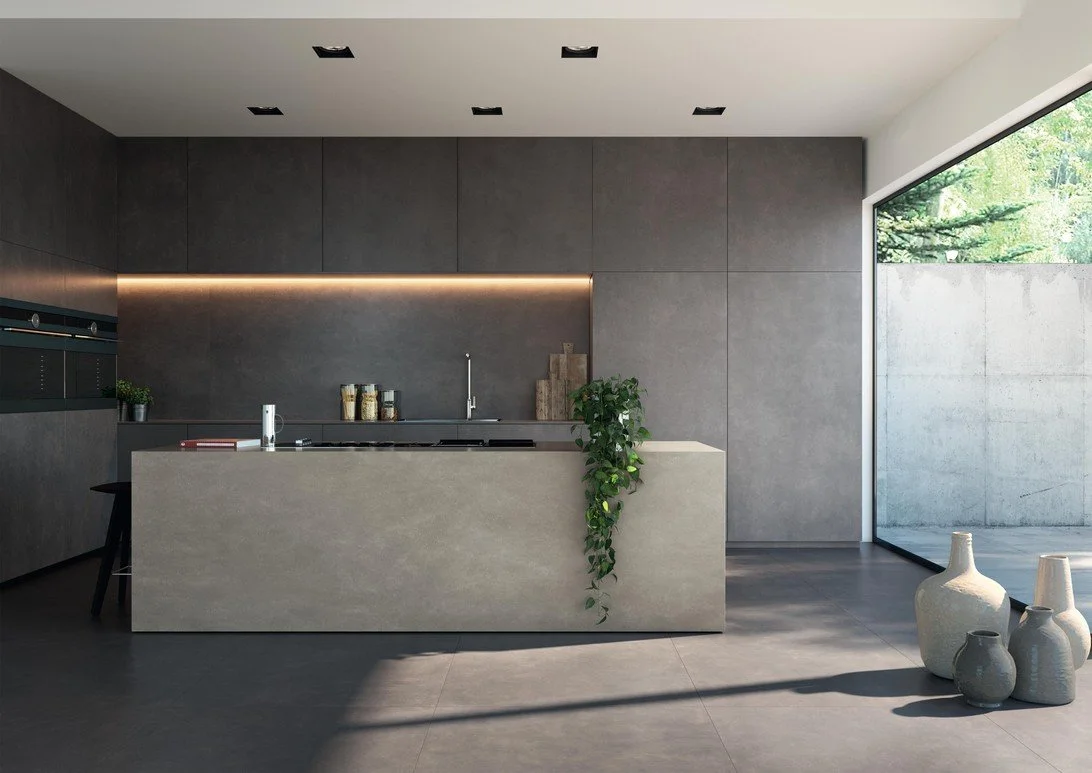 Modern minimalist kitchen with dark cabinets, a white island with a plant, and large floor-to-ceiling window showing outdoor greenery . Florim Ceramiche Biotech