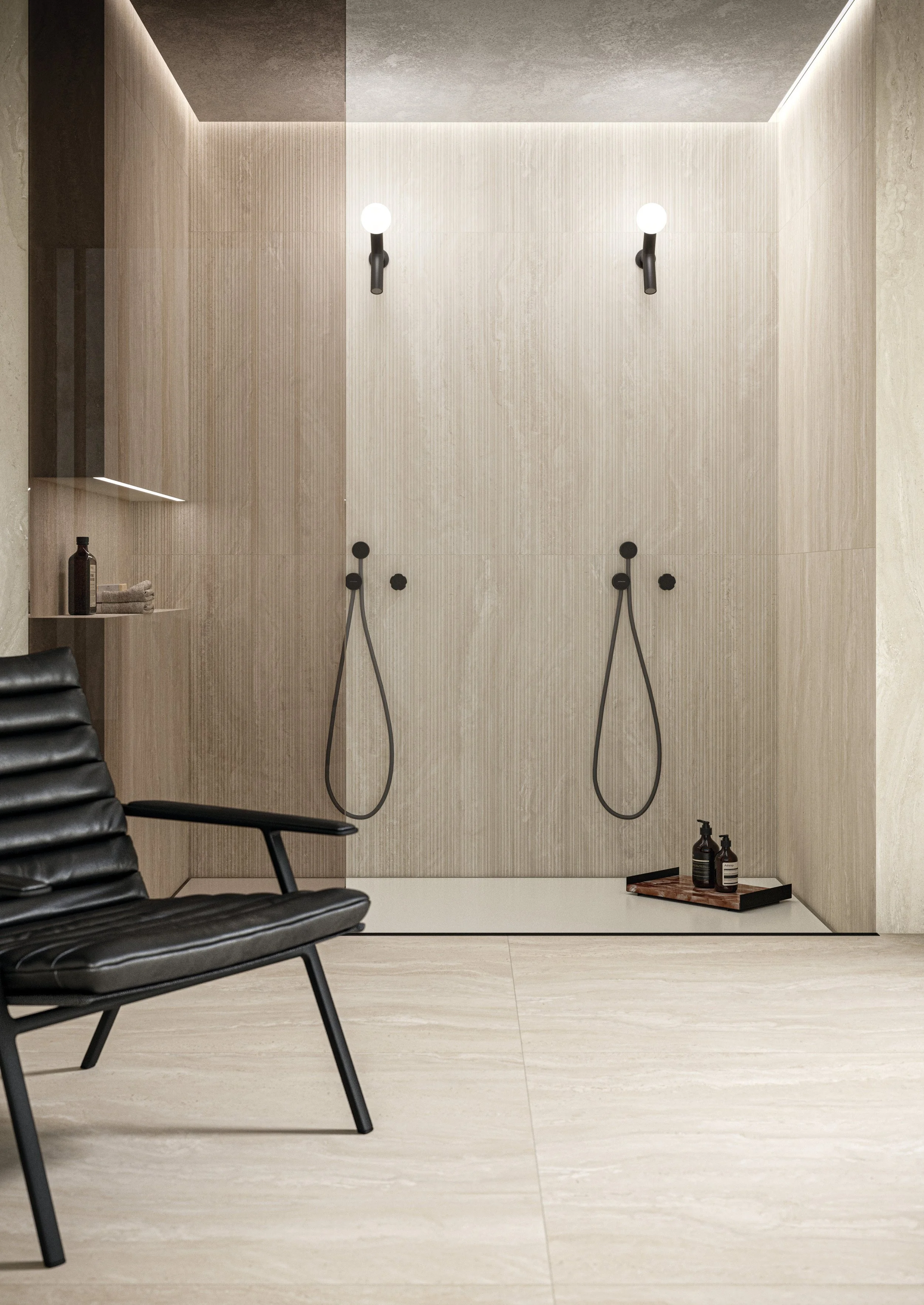 Modern shower area with two handheld showers, black fixtures, bottles and a tray on the floor, in a minimalistic bathroom with beige walls and light-colored tiles.  Florim Ceramiche Authentic Luxe