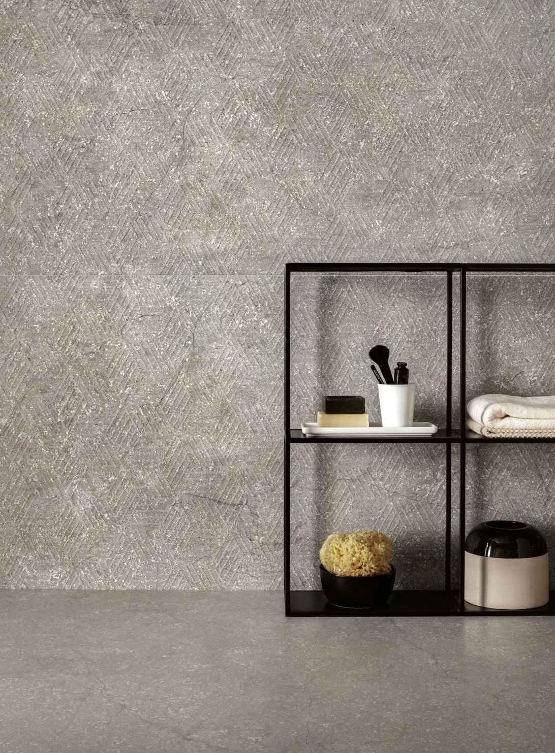 A black metal storage shelf holding soap, a sponge, towels, and a small appliance, against a textured gray wall. Florim Ceramiche Stone Life