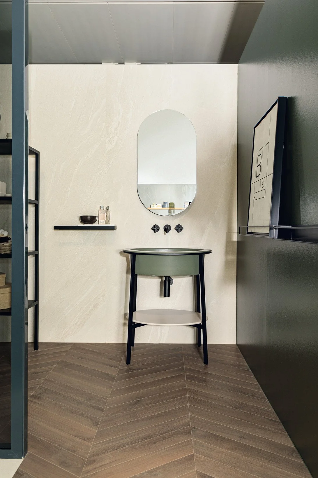 Minimalist bathroom with a black sink, oval mirror, black shelf with toiletries, and a black framed television on a gray wall. Florim Ceramiche Woodlsate Life