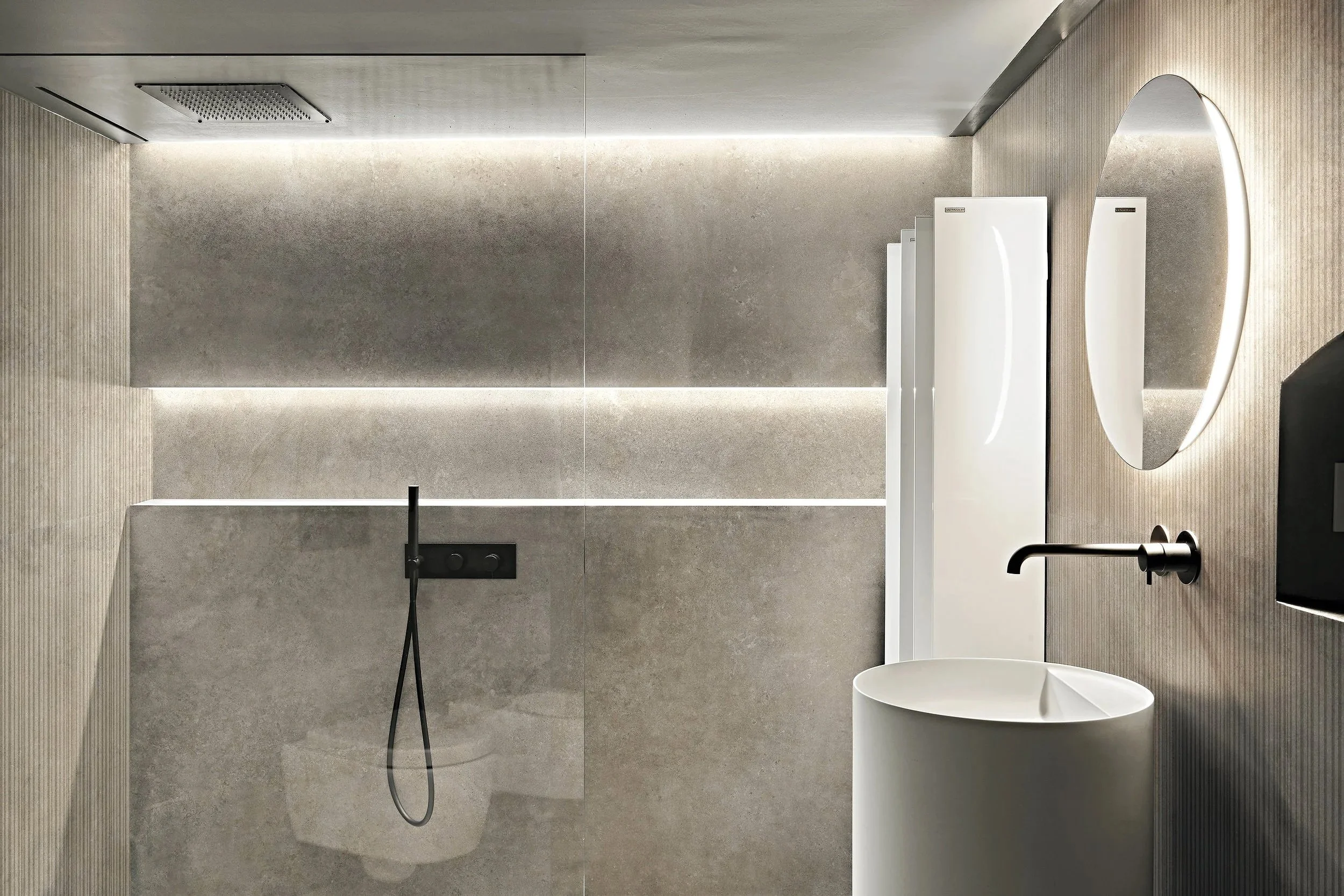 A modern minimalist bathroom with a large round mirror, black faucet, white bathtub, and a walk-in shower with a black showerhead and handheld shower. Florim ceramiche , Casual Life