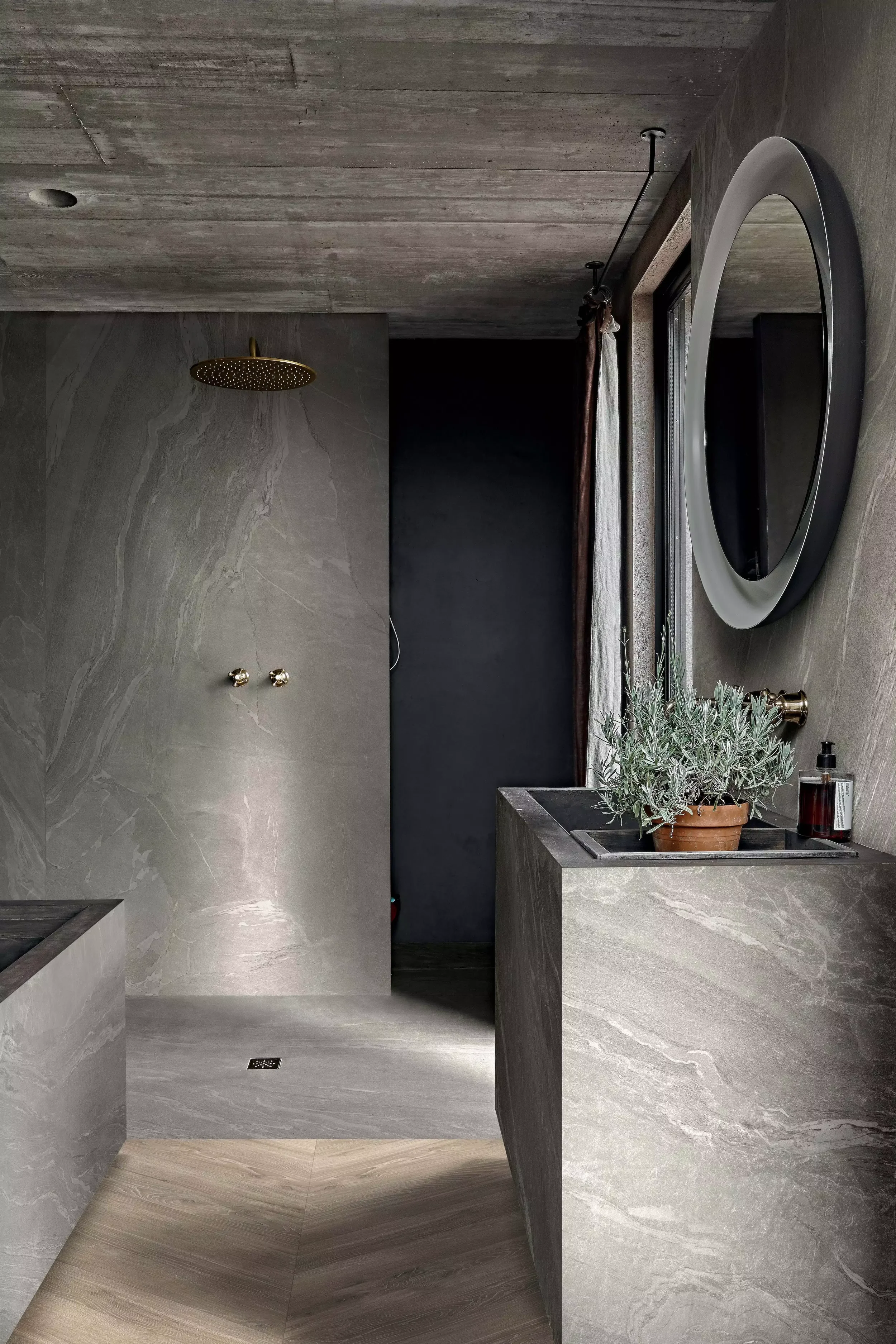 Modern bathroom with gray marble walls and floors, a round mirror on the right wall, a window with curtains, a green potted plant in a brown pot on the sink counter, and a bottle of soap or lotion. Florim Ceramiche Woodslate Life