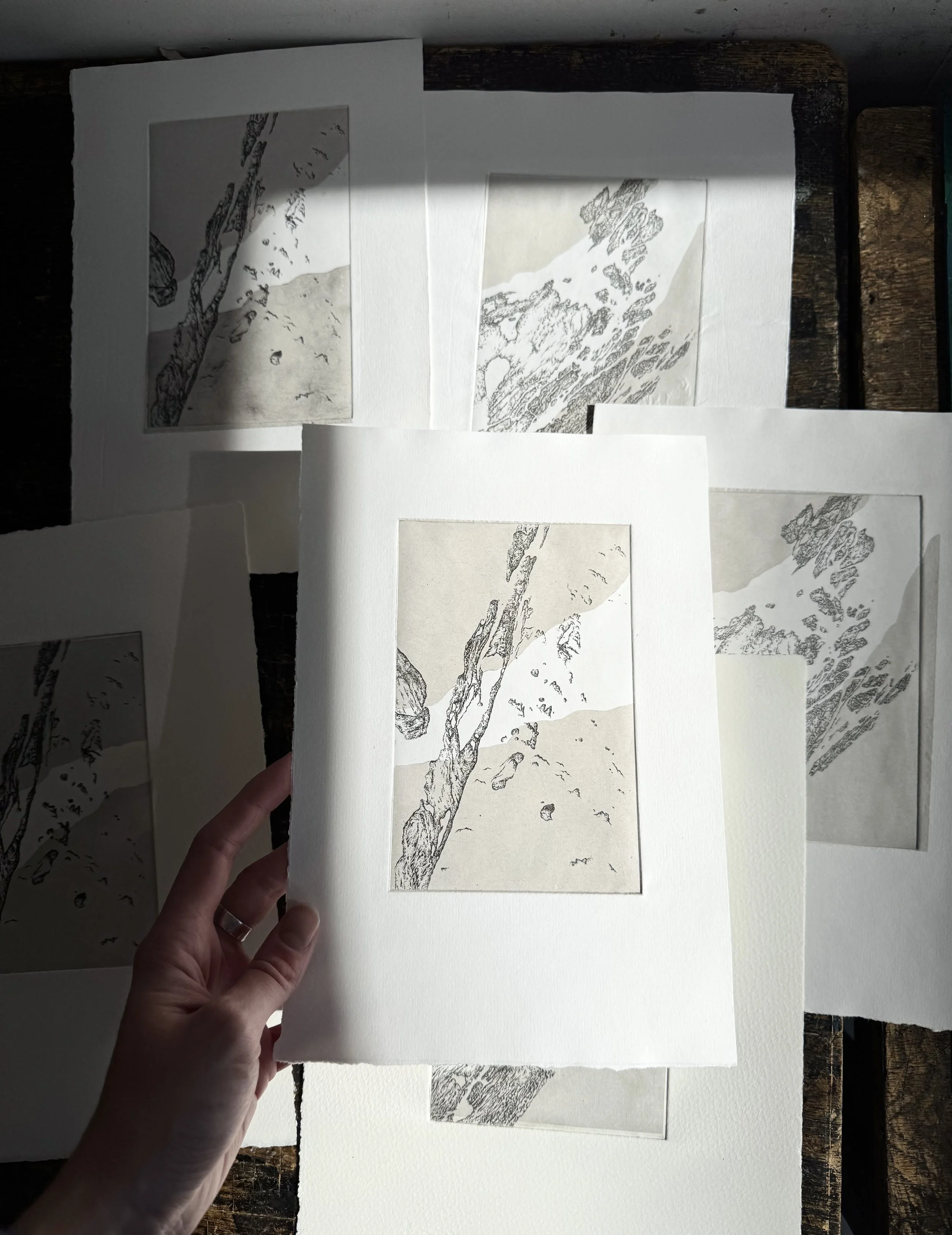 Hand holding a sheet with a black and white sketch of a tree and mountains, placed on a white mat surrounded by other sheets with similar sketches.