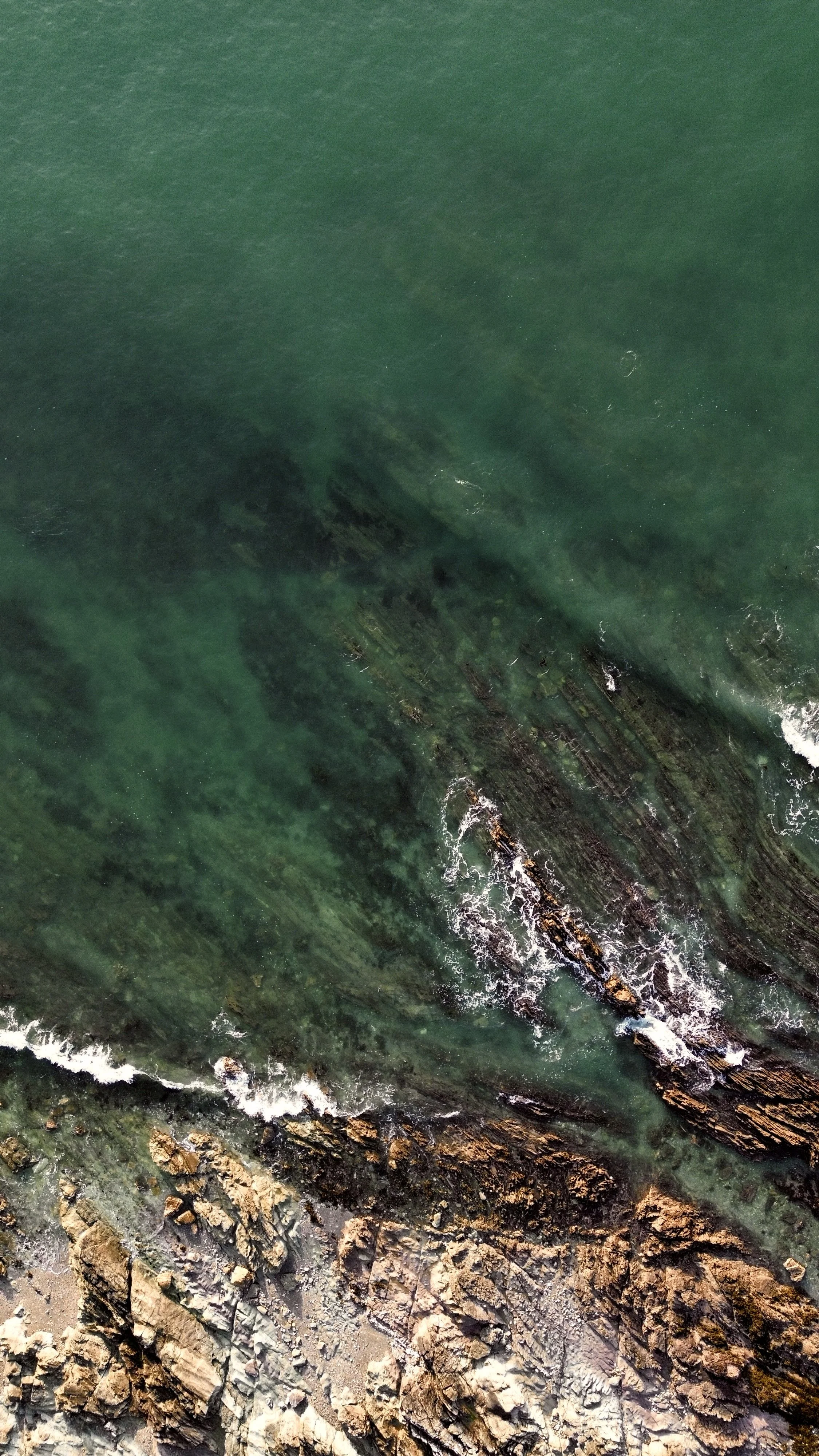 An aerial view of a rocky shoreline with clear green water and waves crashing against the rocks.