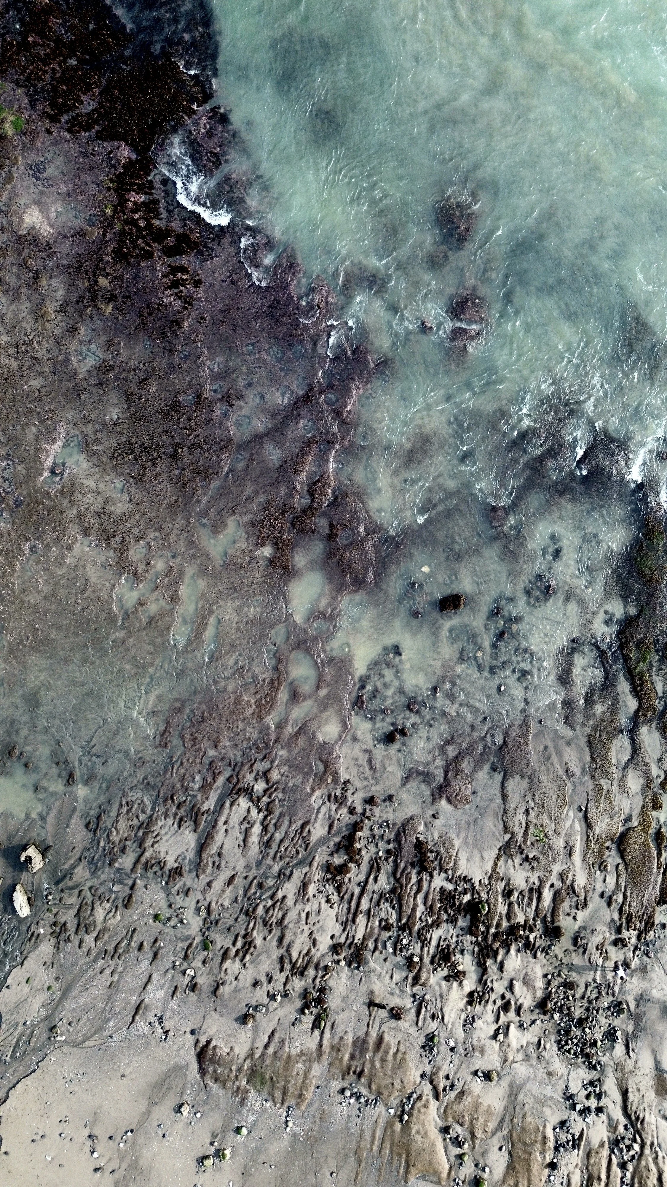 Aerial view of a rocky shoreline with water washing over rocks and sandy beach area.