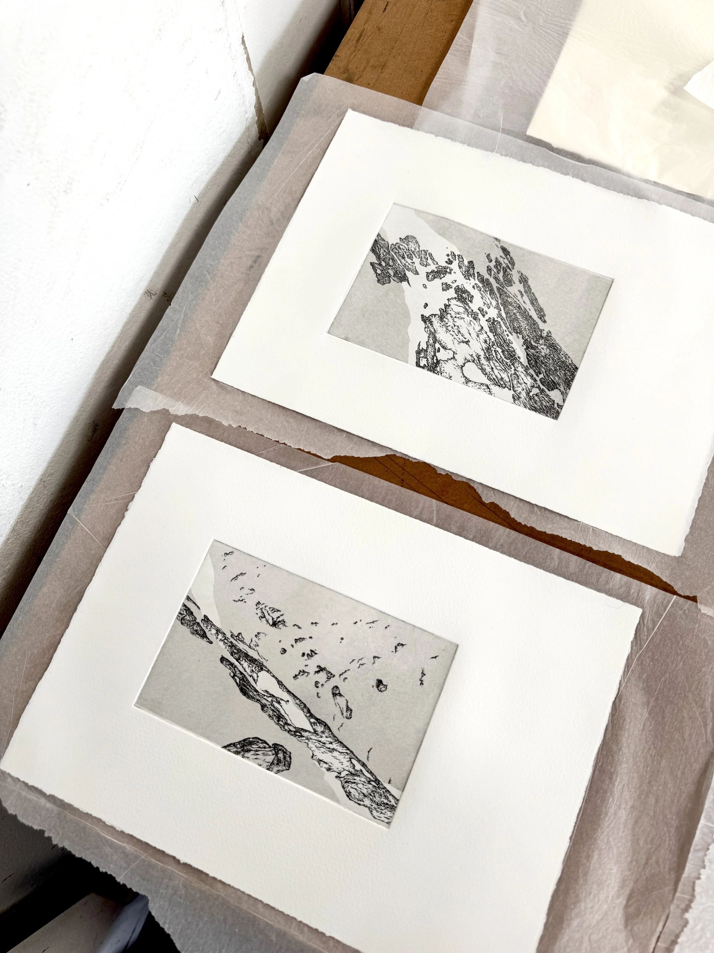 A collection of black and white landscape prints laid out on a white surface, with some framed and others unframed, partially covered with tissue paper.