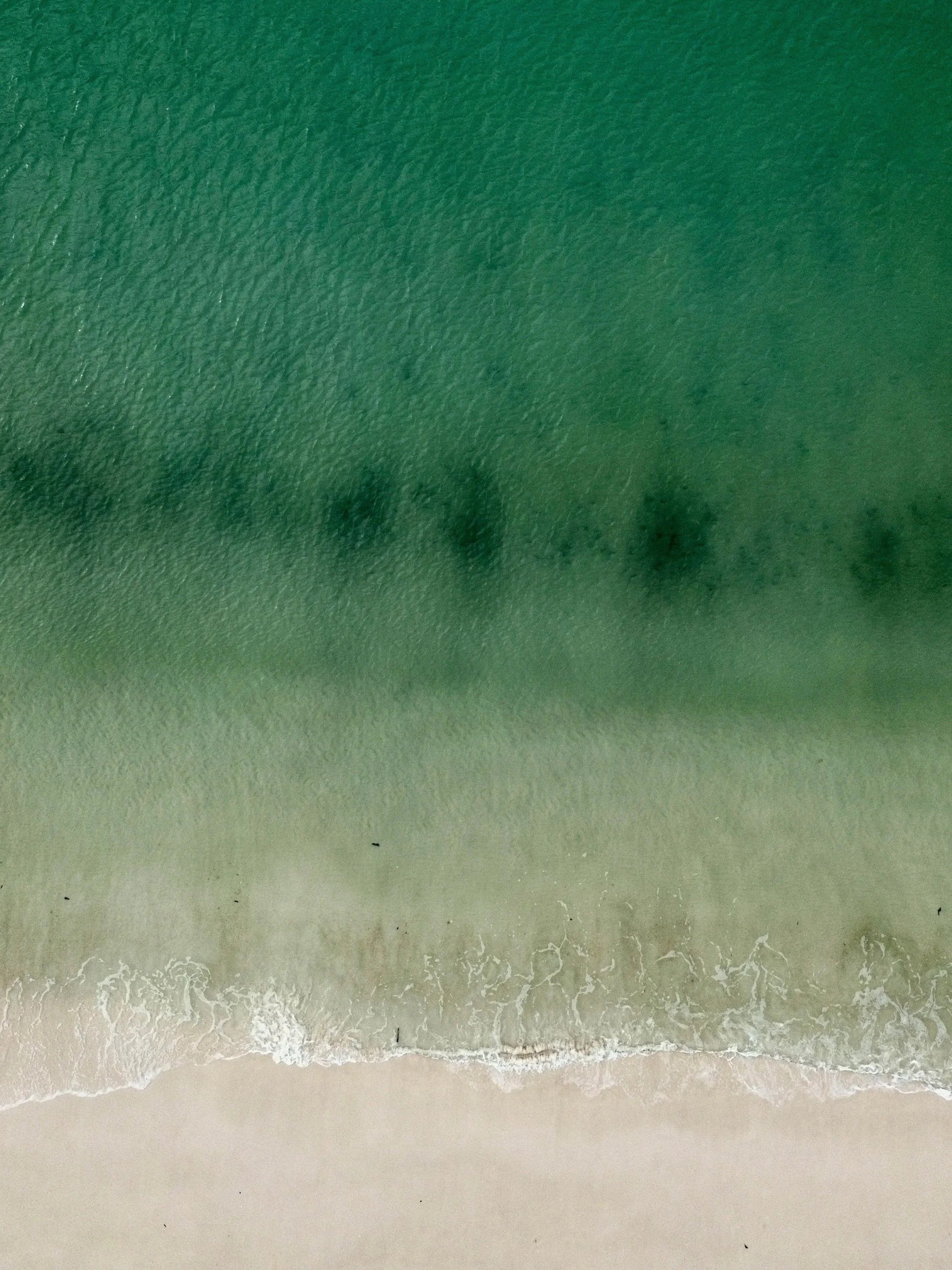 Aerial view of a sandy beach with gentle waves washing ashore, transitioning from deep green in the water to light beige on the sand.