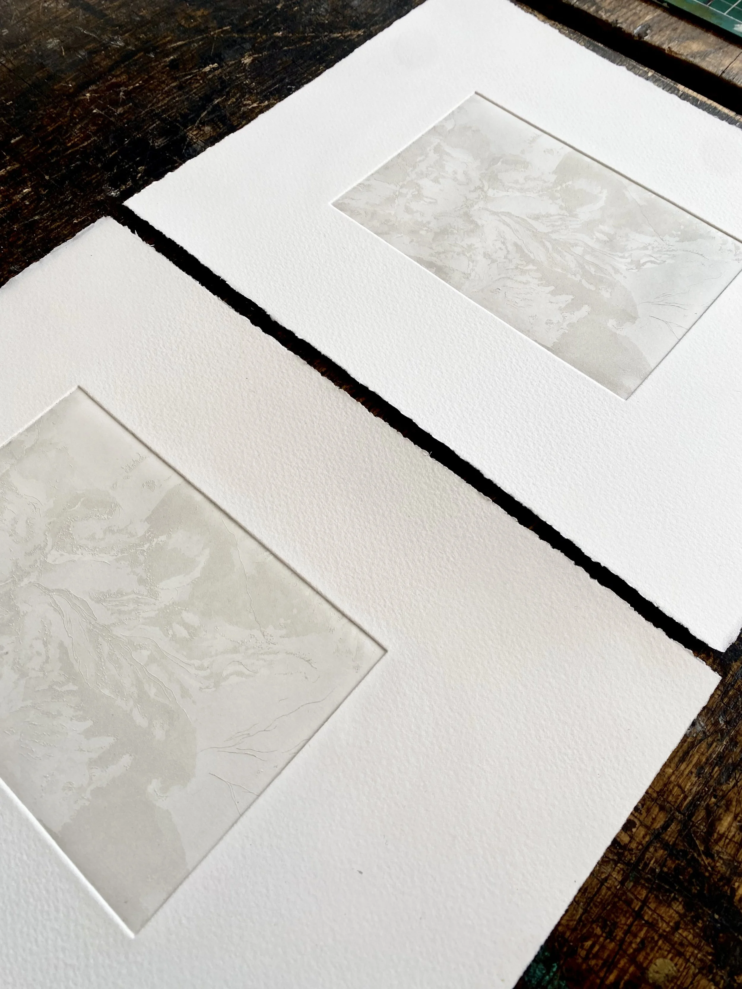 Close-up of a sheet of textured white paper with two transparent marbled paper samples placed on it, on a dark wooden surface.