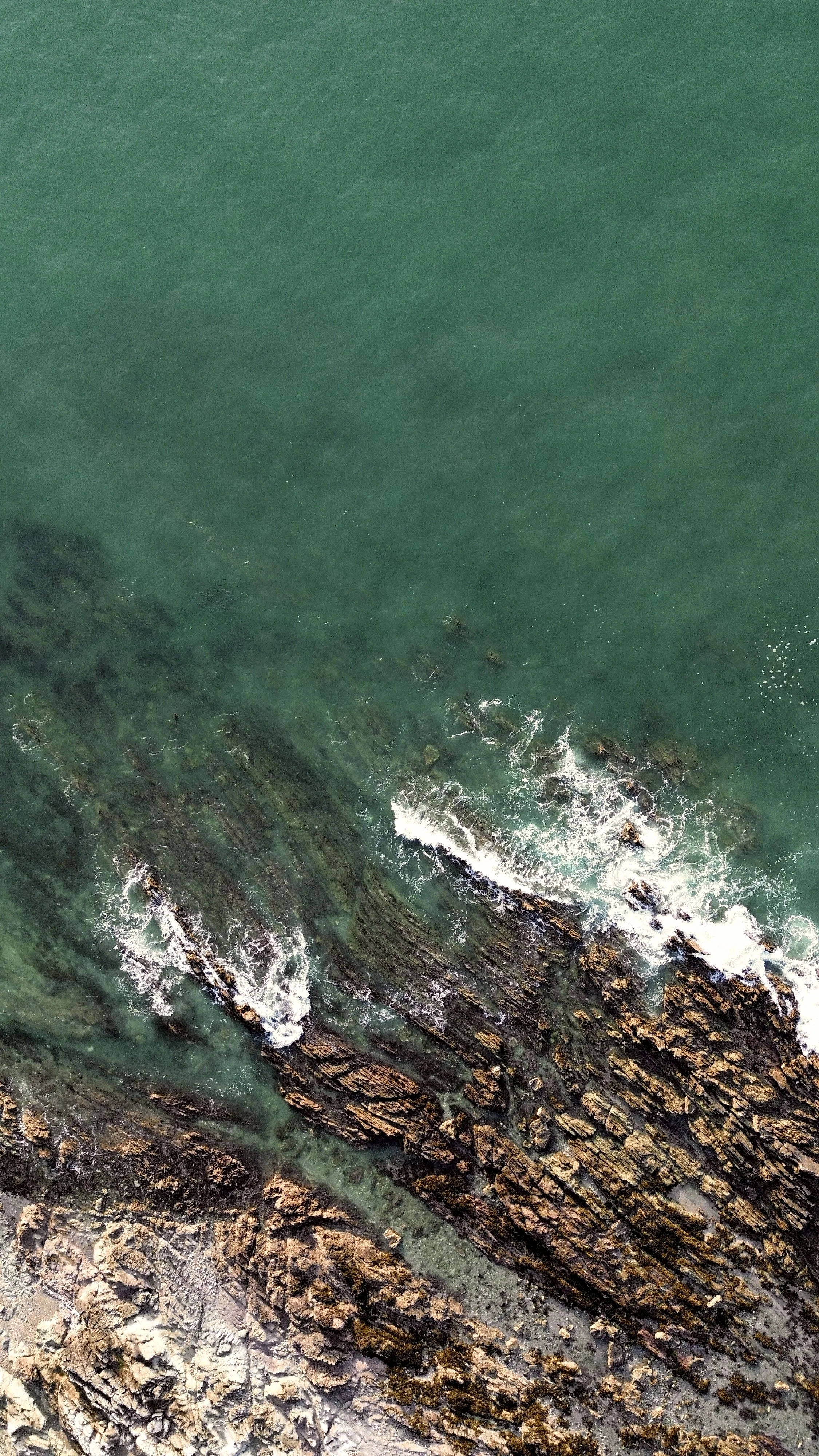 Aerial view of ocean waves crashing against rocky shoreline with visible underwater rocks.