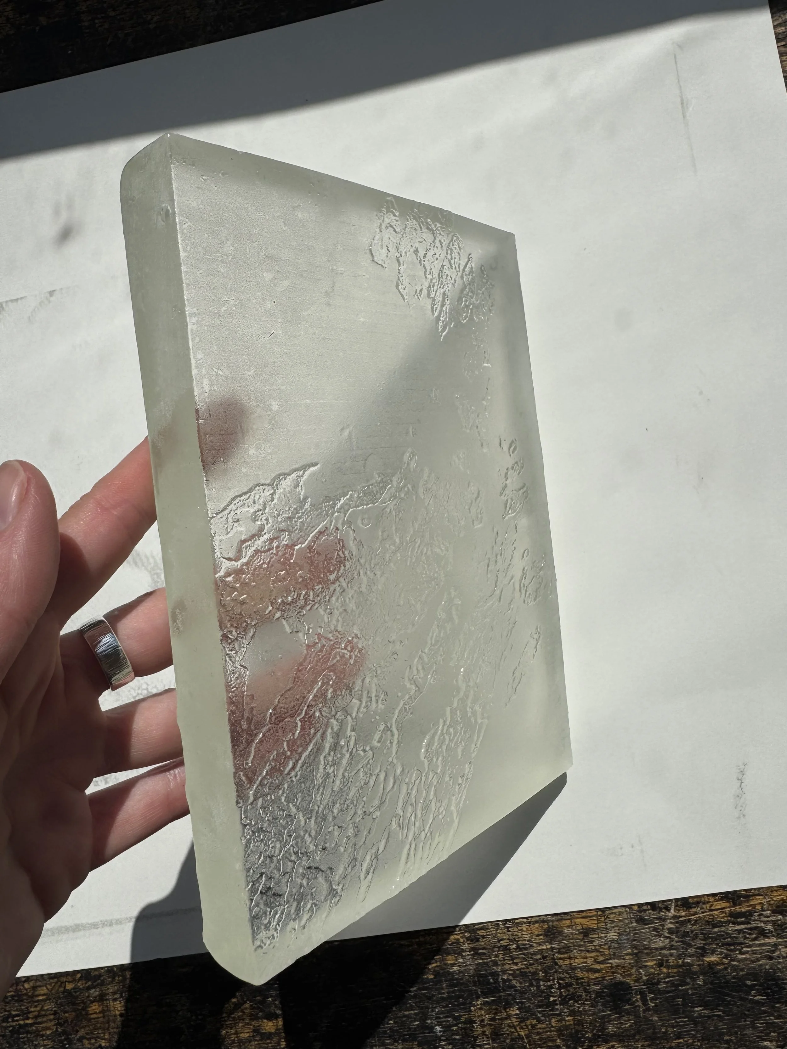 A hand holding a transparent, textured glass sheet with a frosted map of the world, slightly angled and casting shadows on a white background.