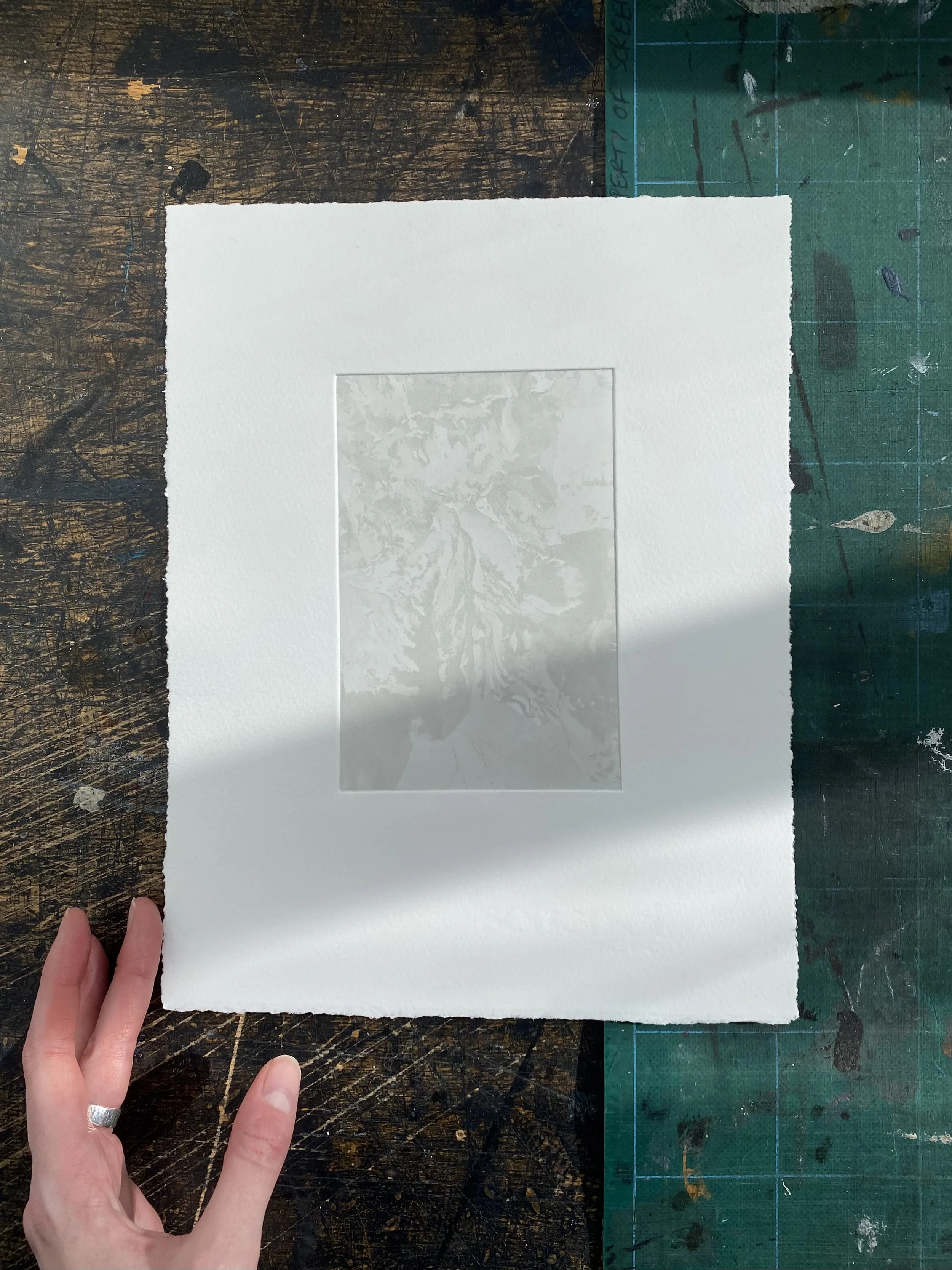 A hand holding a piece of white textured paper with a transparent rectangular window showing a reflection of trees and sky, on a dark, worn wooden surface with a green cutting mat nearby.