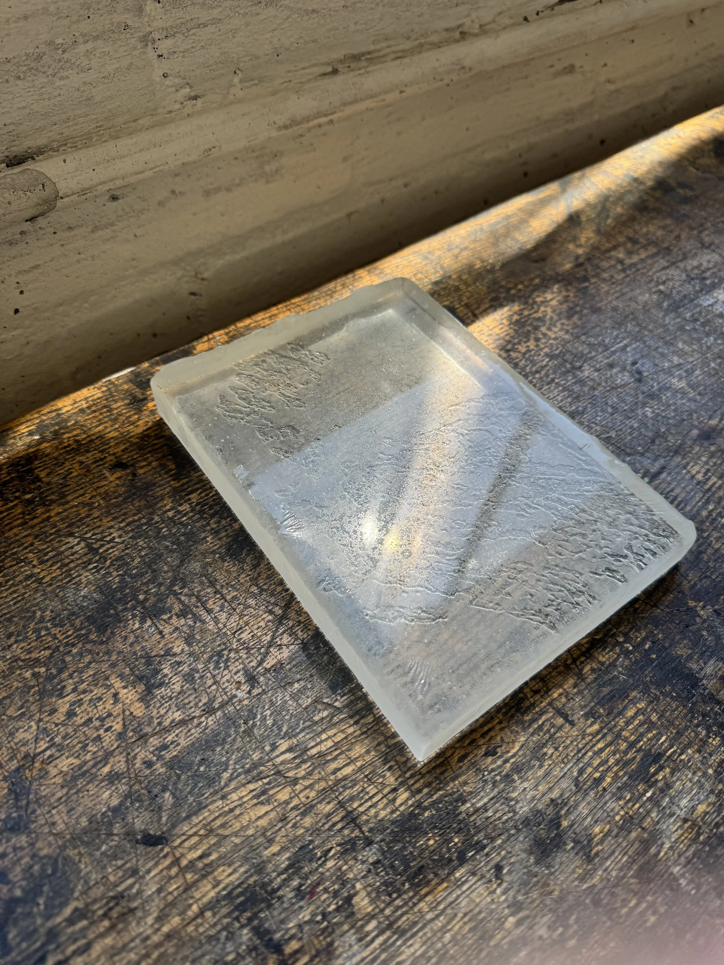 A clear rectangular ice cube tray partially filled with water, resting on a dark, scratched wooden surface, with a background of a stained and worn wooden wall.