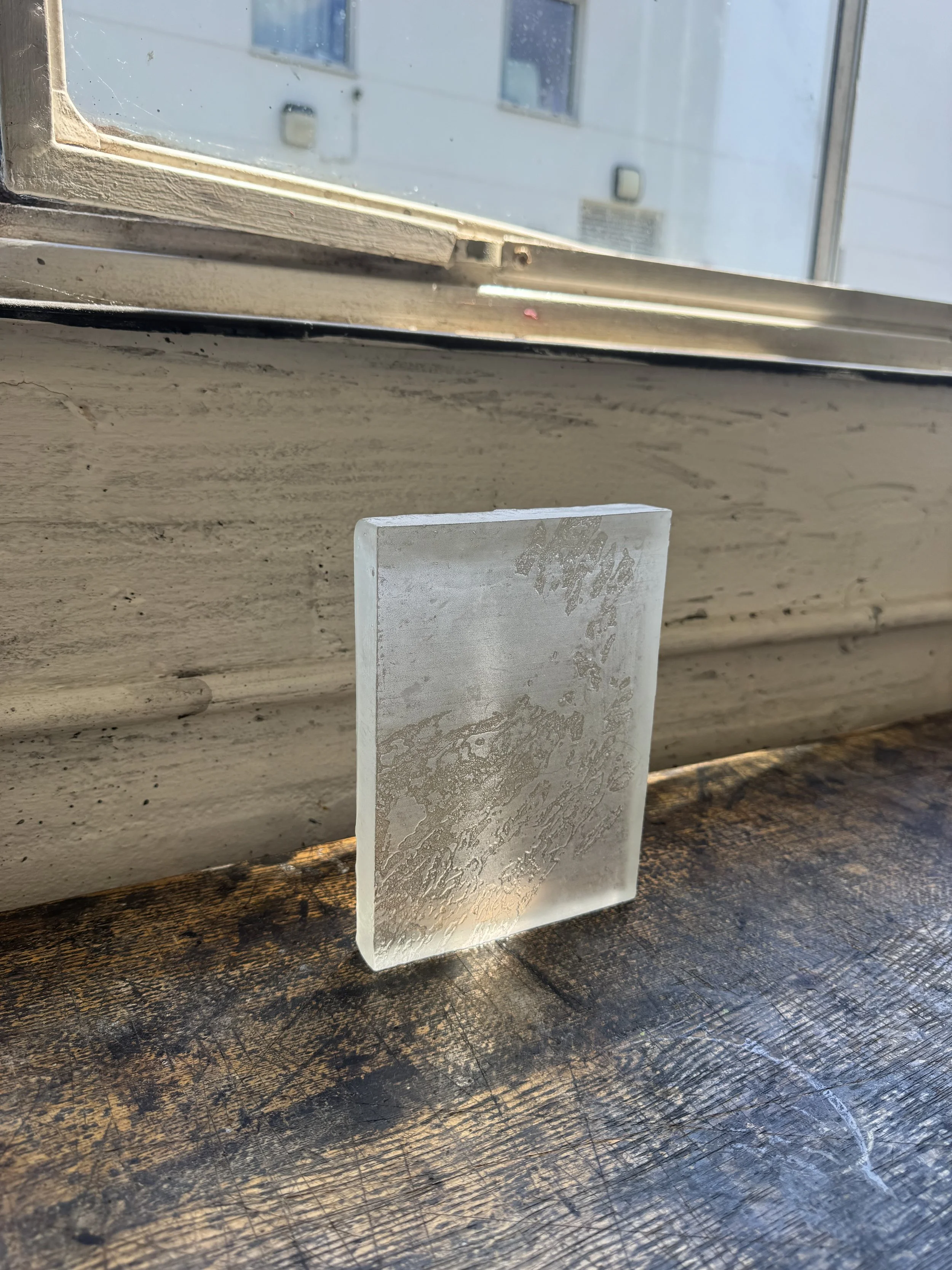 A small piece of translucent, frosted glass with a textured surface, leaning against a worn, dark wooden surface. The background includes a windowsill and a window with buildings outside.
