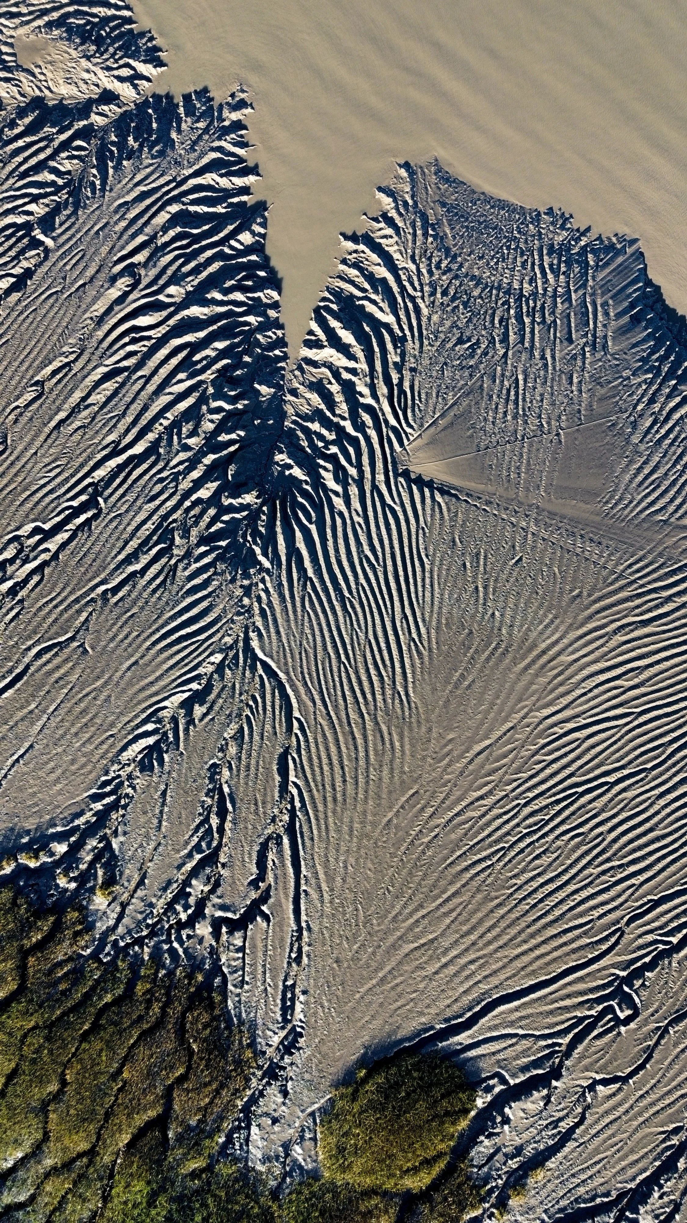 Aerial view of sand dunes with intricate patterns and some green vegetation at the bottom.