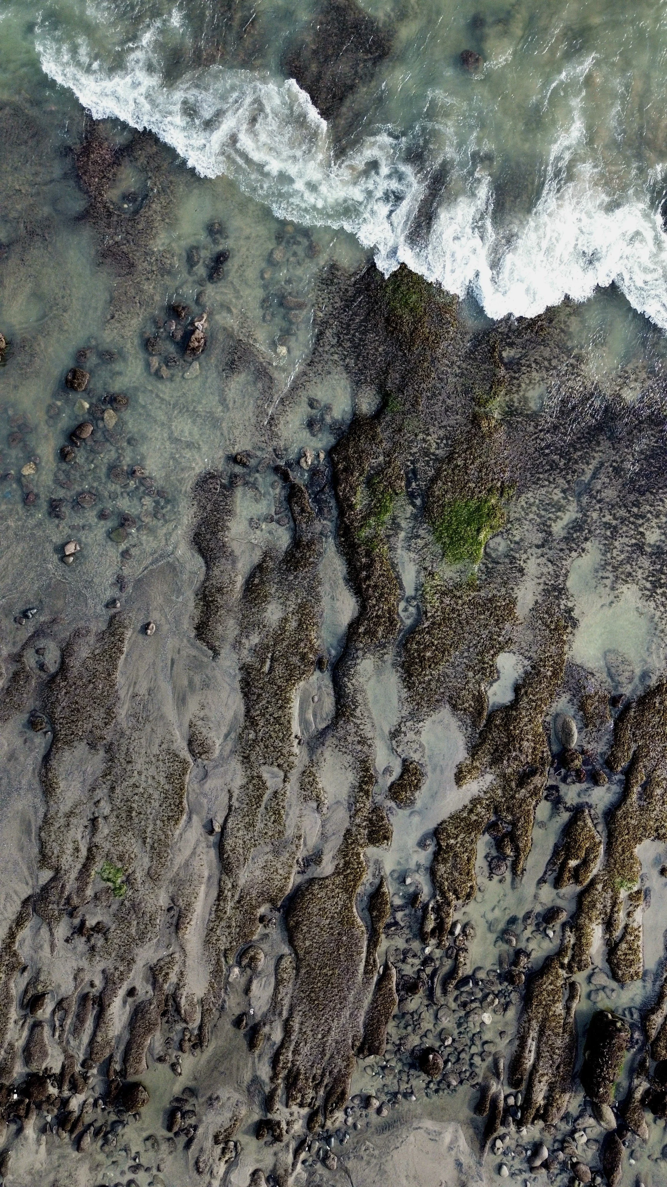 Aerial view of a shoreline with rocks and waves washing over the sand and rocks, creating patterns in the wet sand.