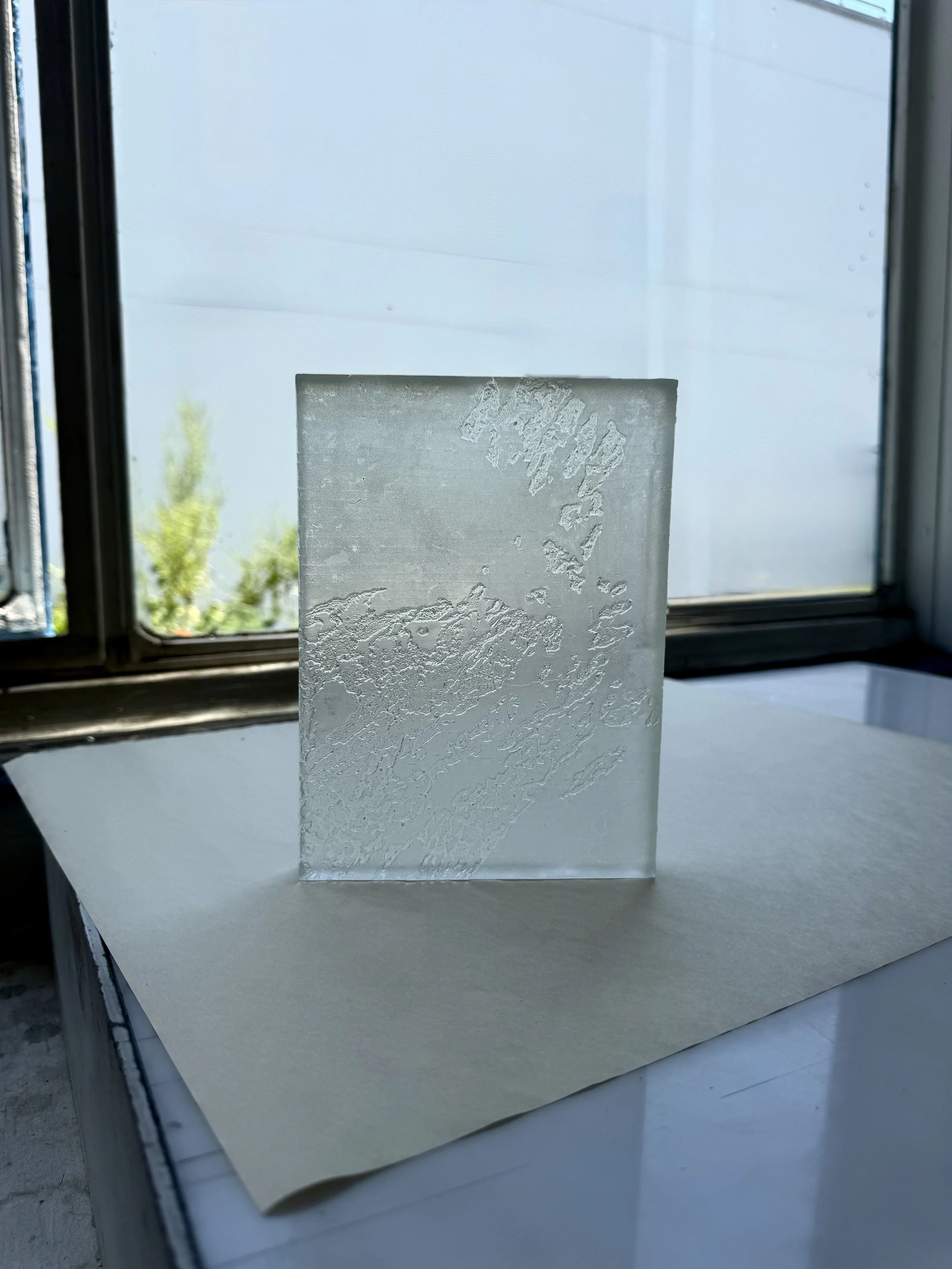 Close-up of a sheet of frosted or textured glass with intricate patterns, placed in front of a window with a view of trees and a pale blue sky.