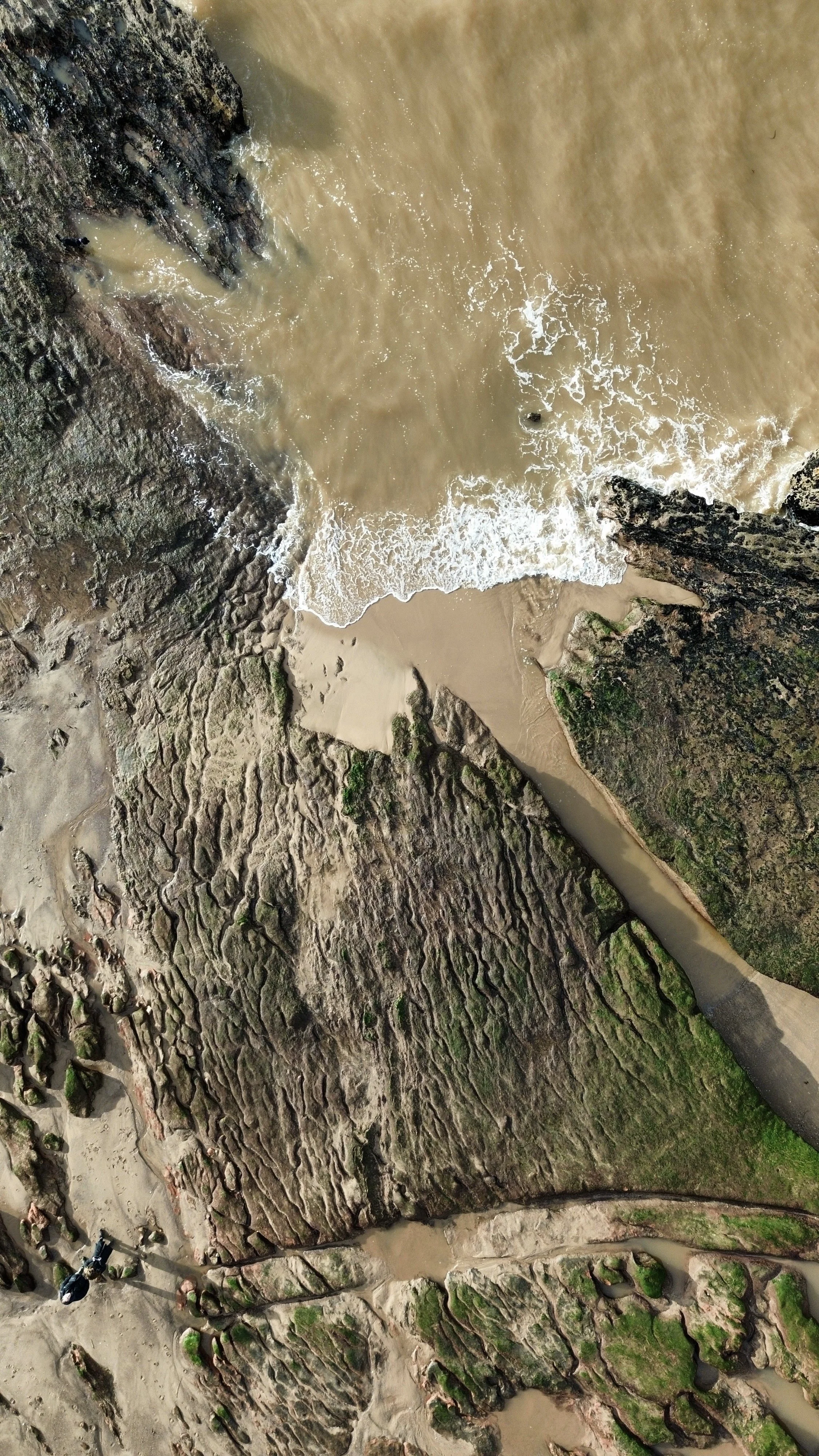 Aerial view of a rocky coastline with waves crashing onto the sandy beach. The rocks are dark and textured with patches of green moss. 