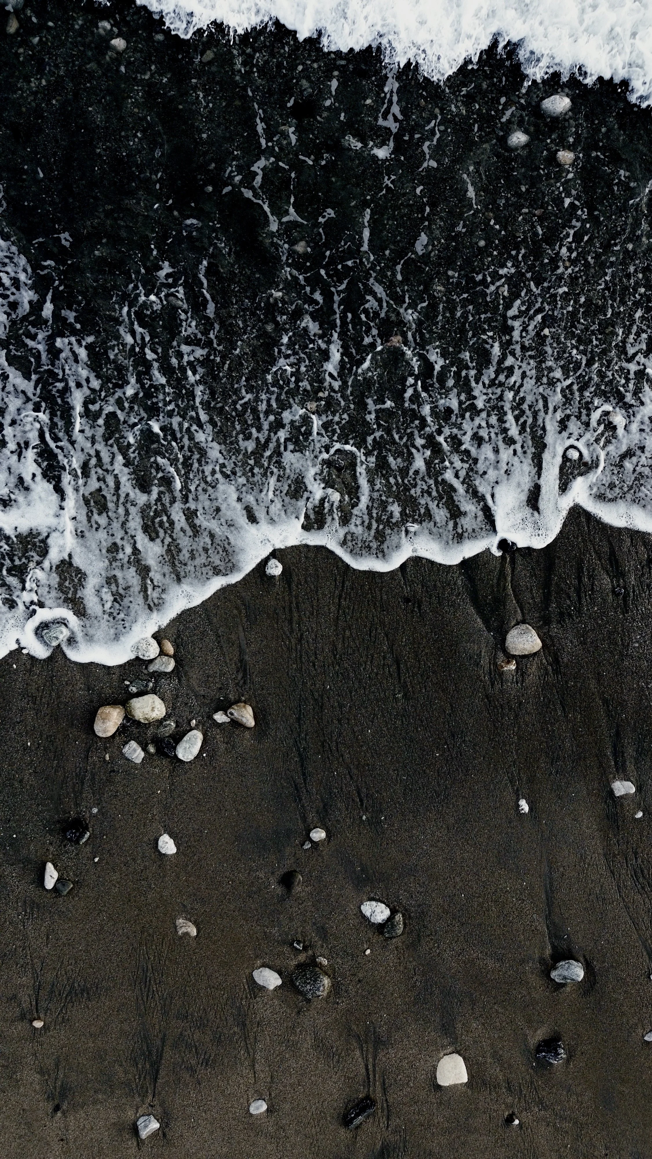 Close-up of black sand beach with small white and black stones, ocean waves washing onto the shore, and foam forming at the water line.