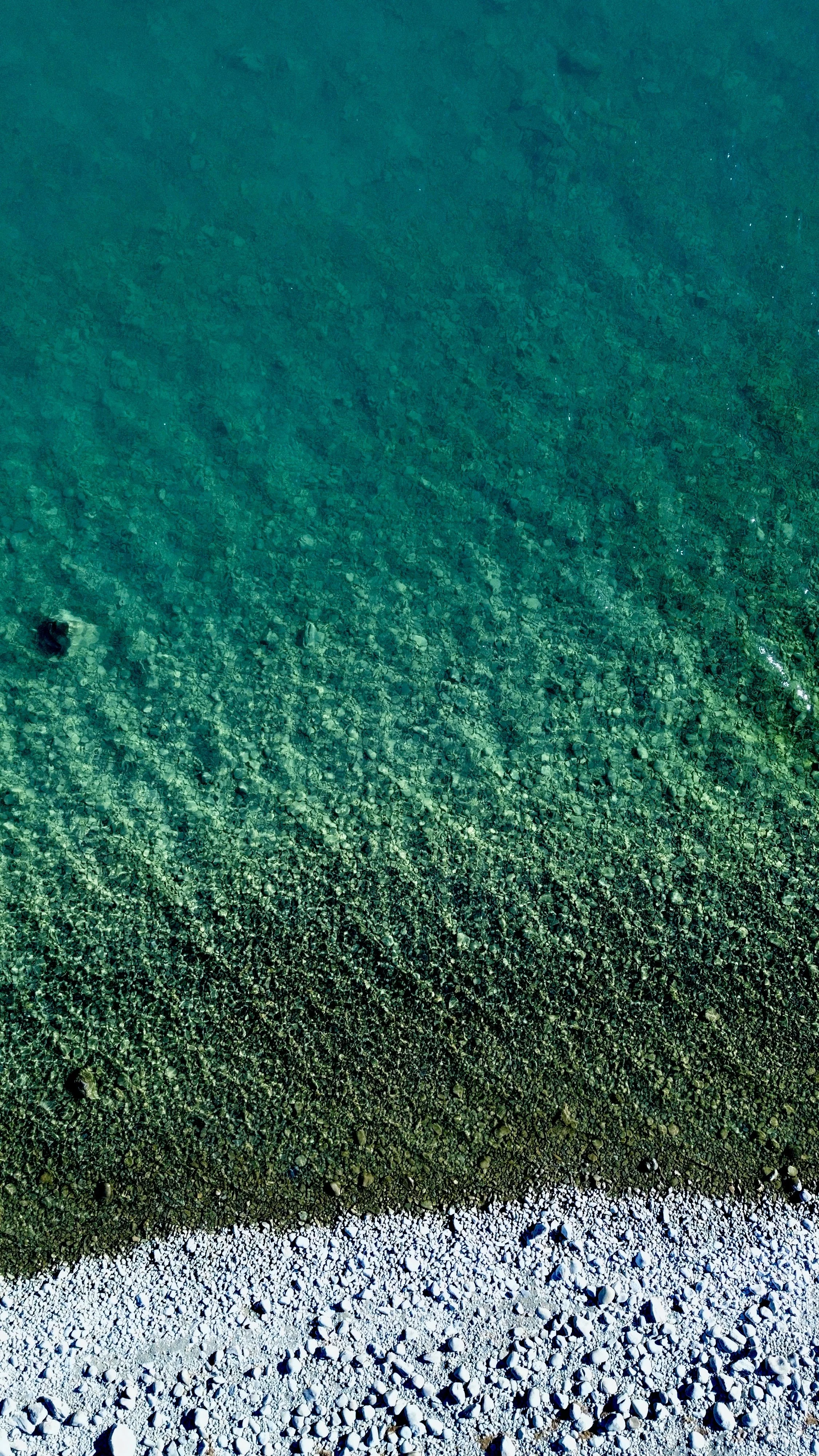 Close-up photograph of a cove showing a gradient of colors from dark green at the bottom, transitioning to teal, and then bluish-green at the top, with visible pebbles and rocks on the bottom.