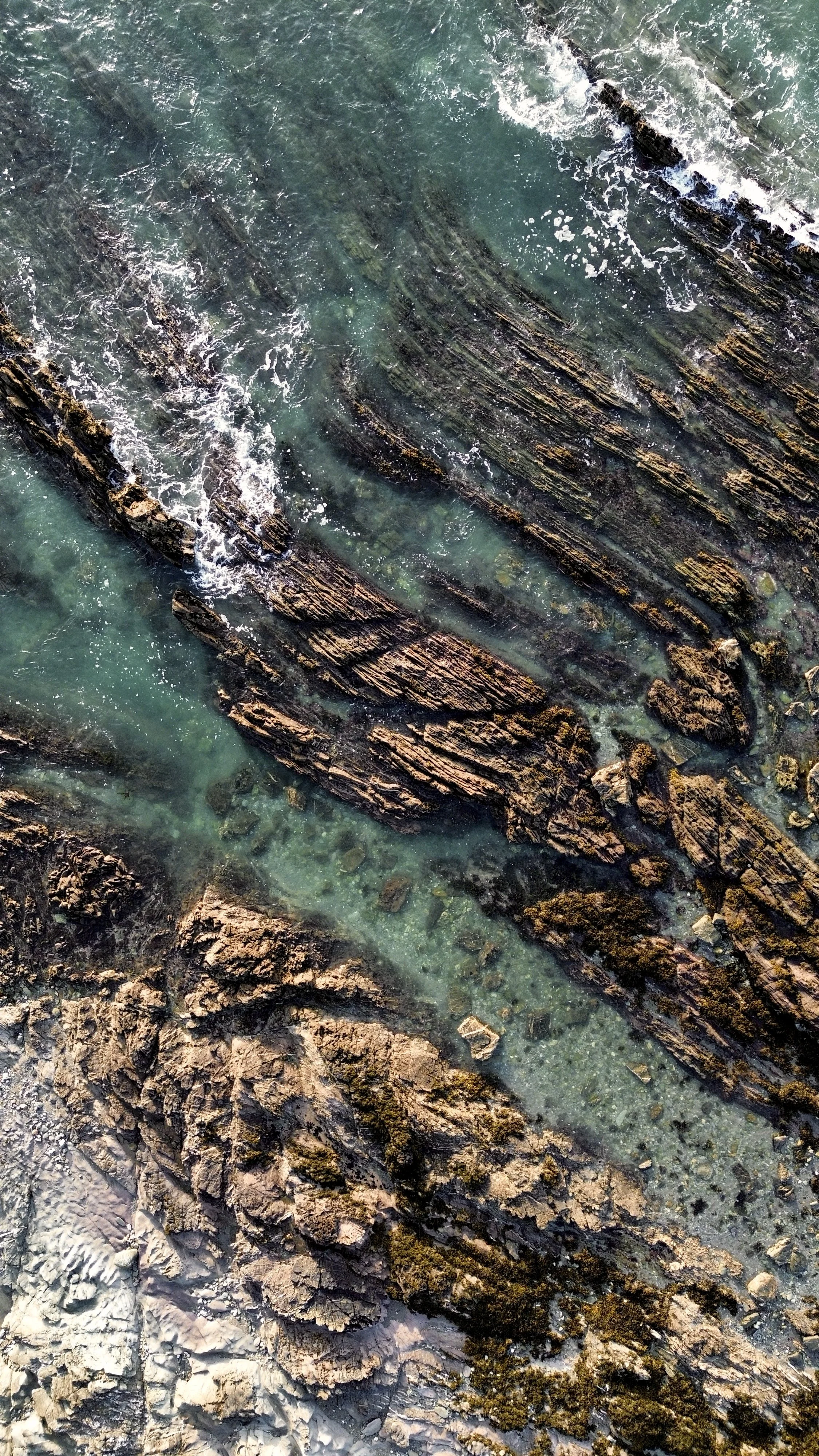 Aerial view of rocky shoreline with layered rocks and clear turquoise water.