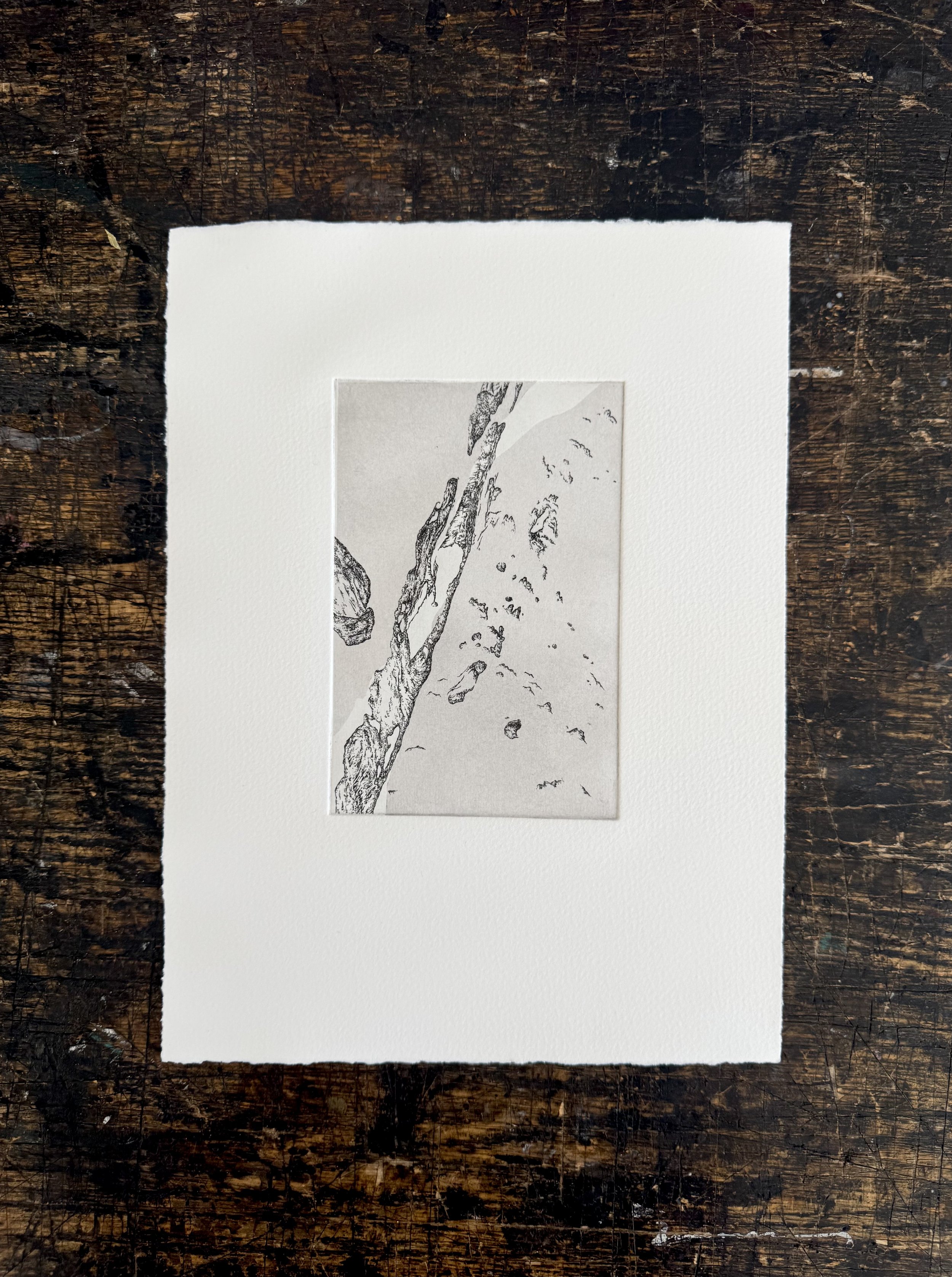 A print of rocks on a piece of paper, framed with white matting, placed on a dark, scratched wooden surface.