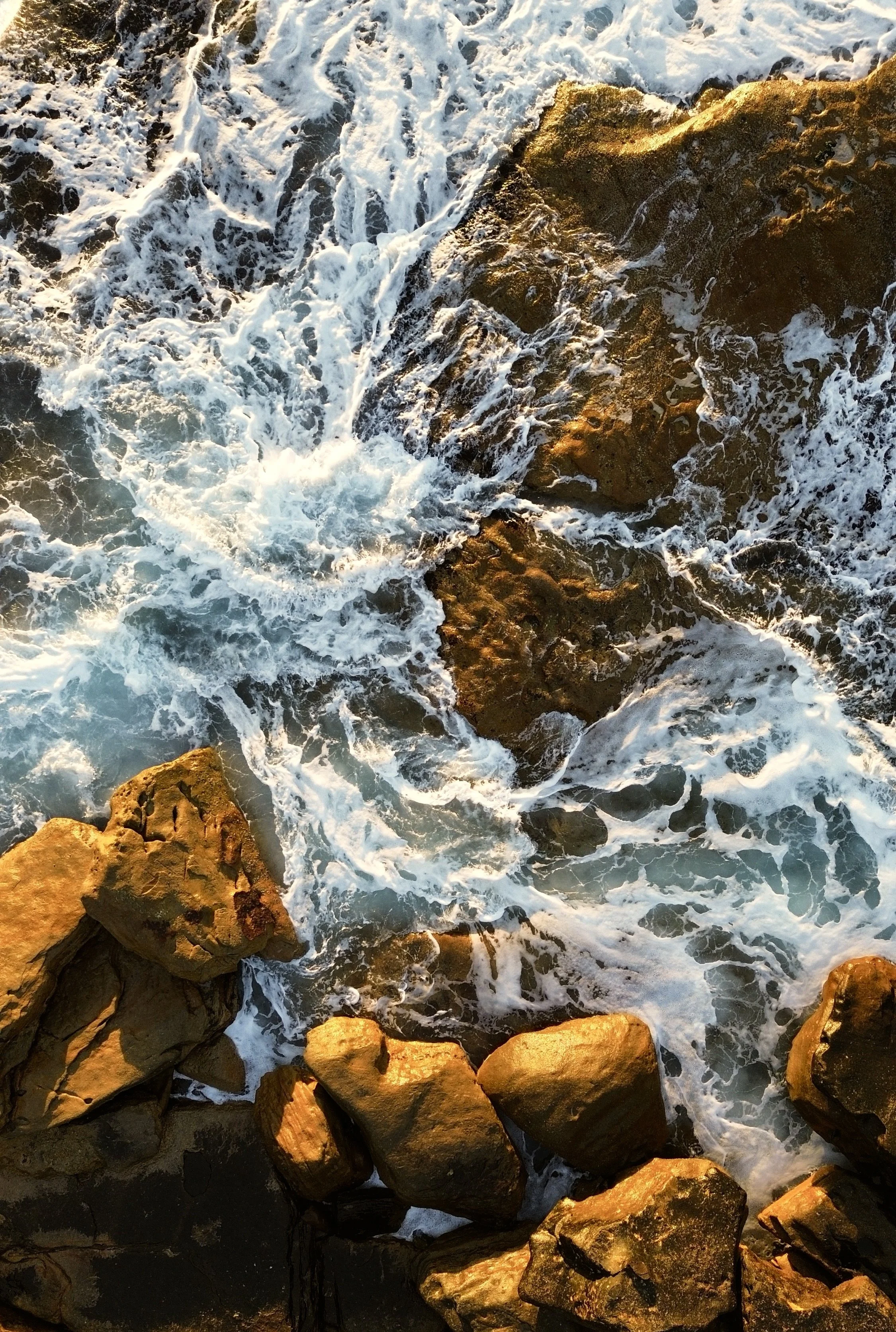 Waves crashing against rocks on the shore, with white foam and some sunlight reflecting on the rocks.