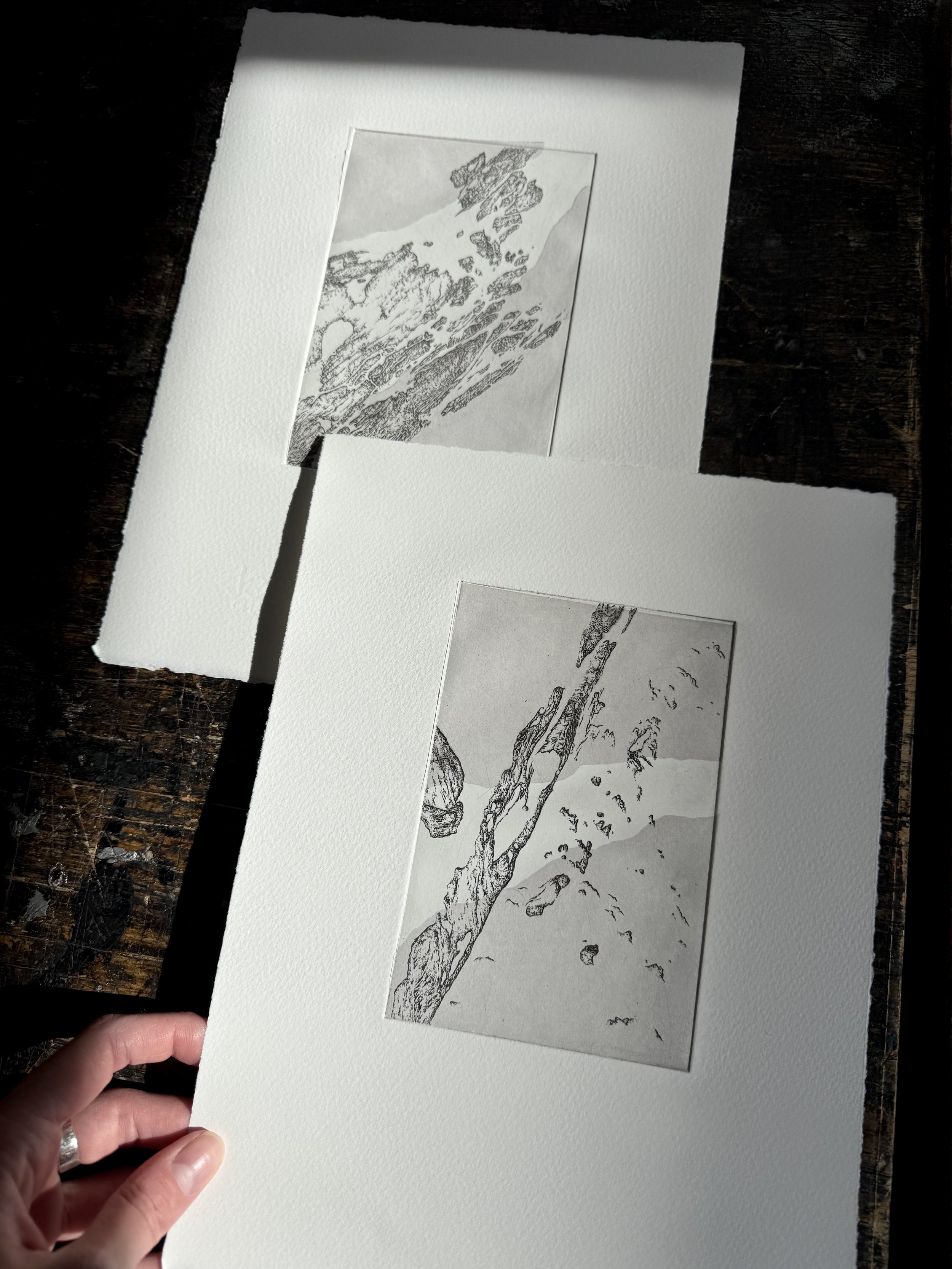 A person holding a white mat with a black and white sketch of a mountain landscape, featuring snow-covered peaks and scattered rocks, on textured paper.