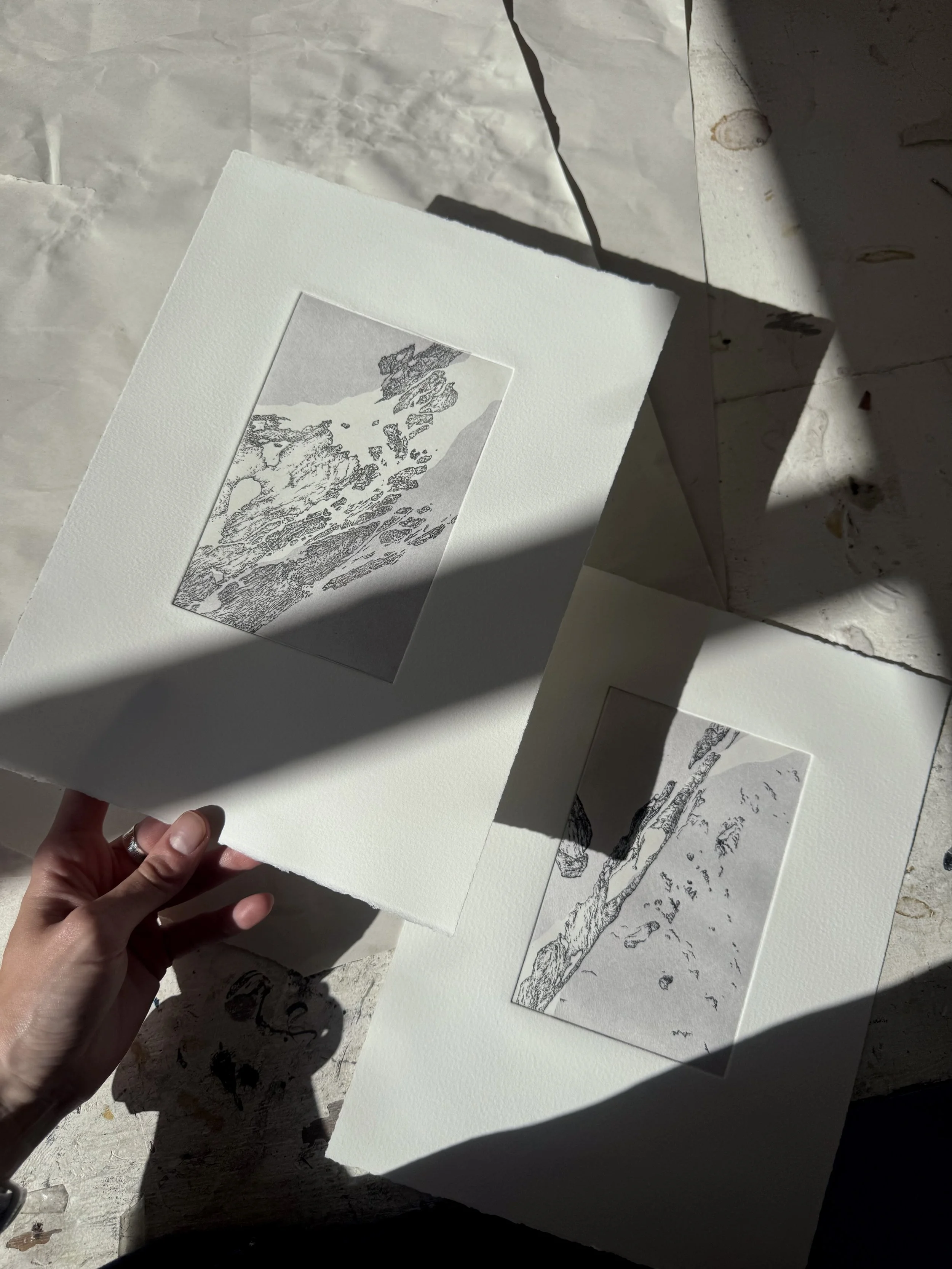 Person holding a sheet of textured white paper with a landscape illustration in black and white. Several other landscape illustrations are spread on a flat surface with sunlight and shadows.