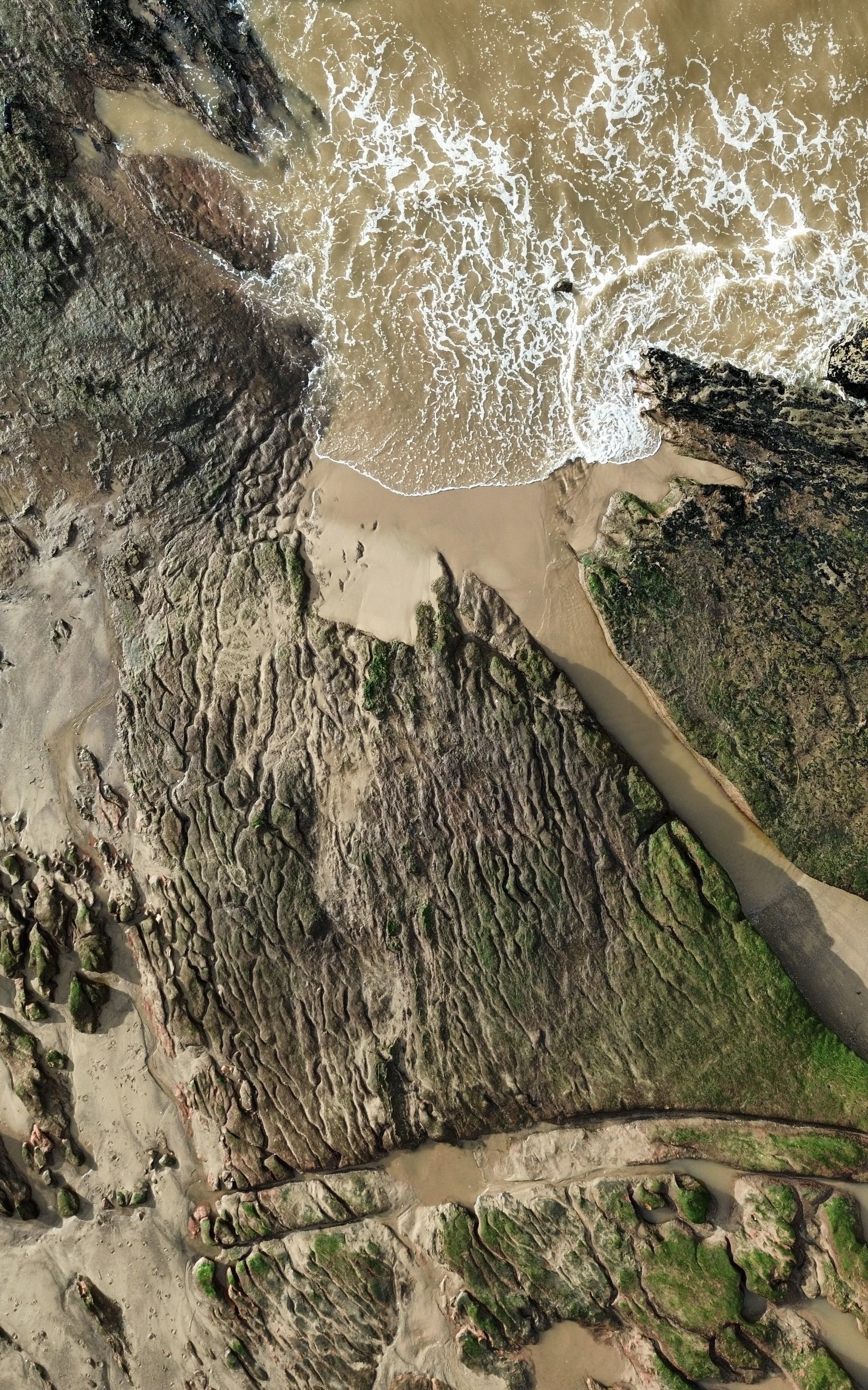 Aerial view of rocky coastline with waves crashing onto sandy beach and green moss on rocks.