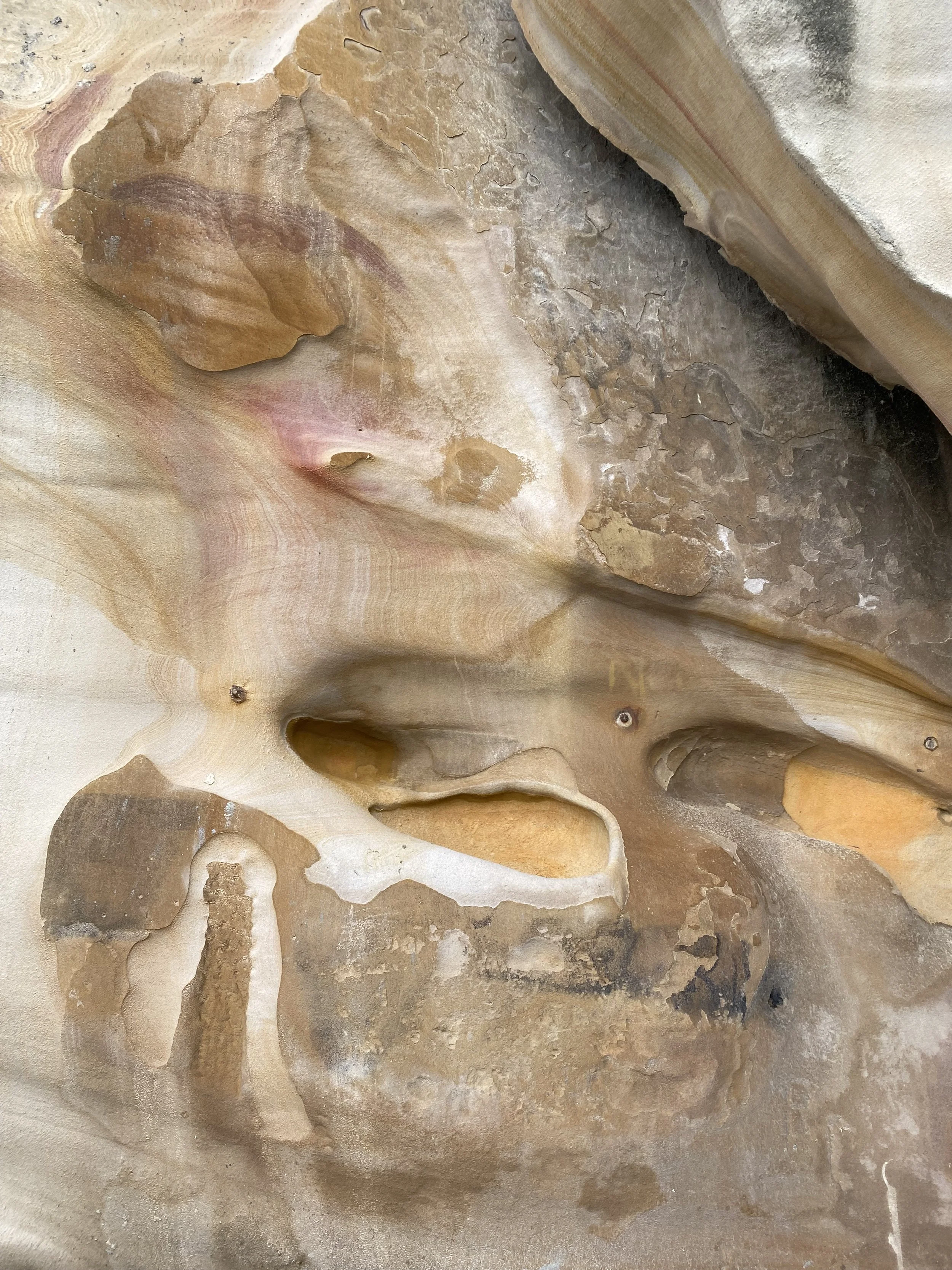 Close-up of layered, weathered rock with various shades of beige, tan, pink, and gray, showing textured and smooth surfaces with irregular patterns and natural formations.