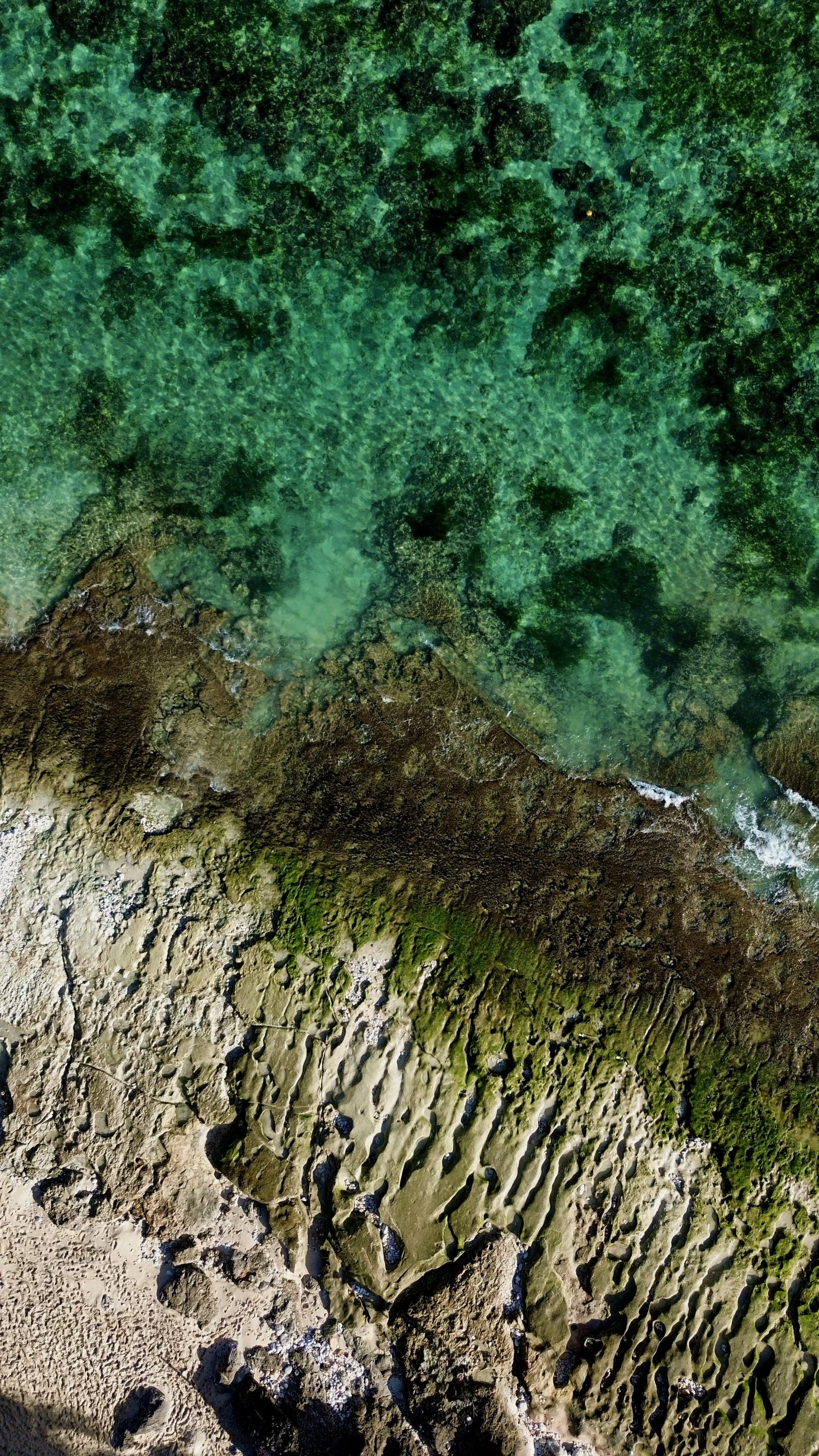 Aerial view of a coastal area with clear green water, rocky shores, and sand with ripple patterns.