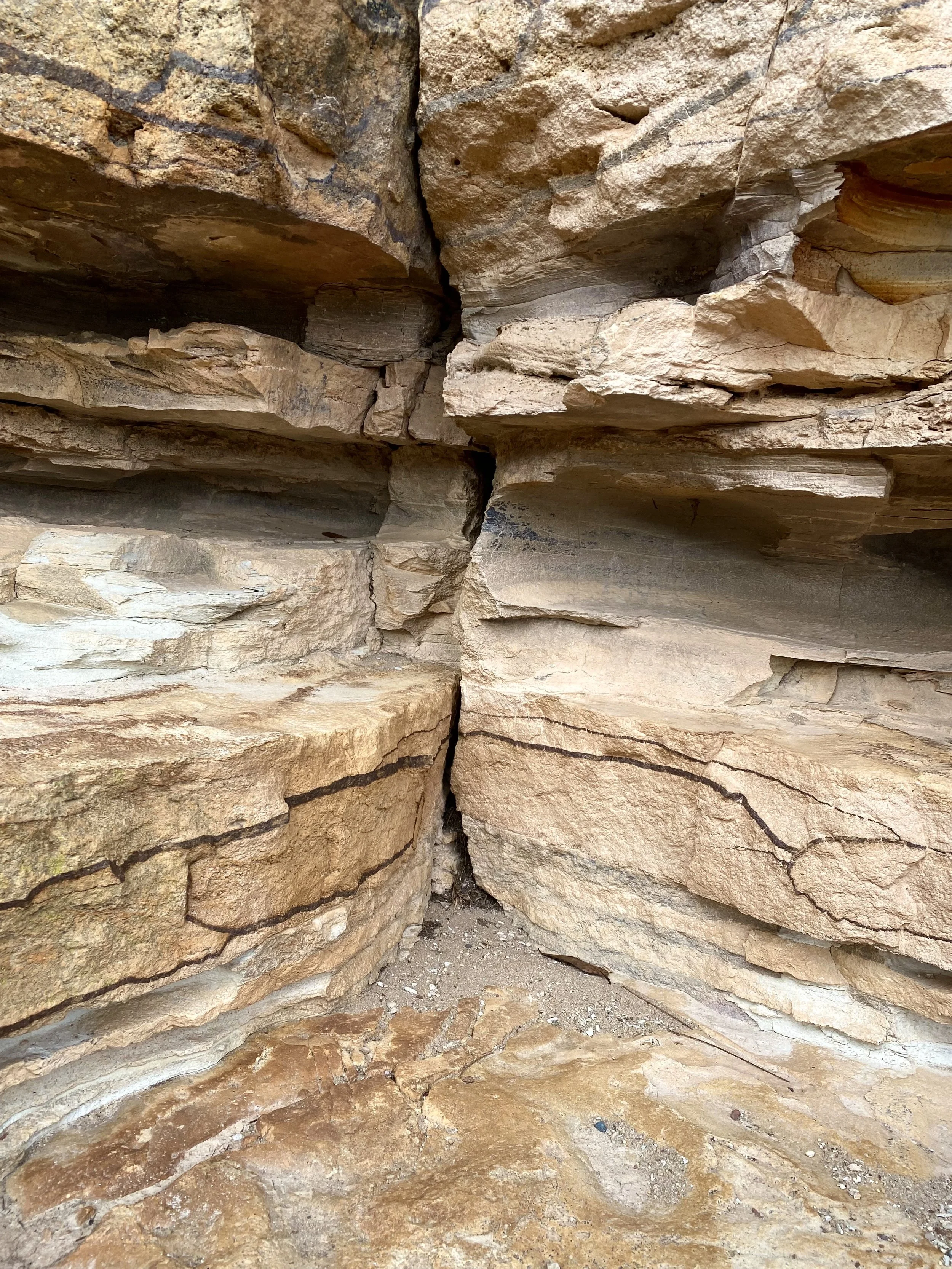 A narrow crevice in a large rocky formation with layered sedimentary rocks in shades of tan, beige, and brown.