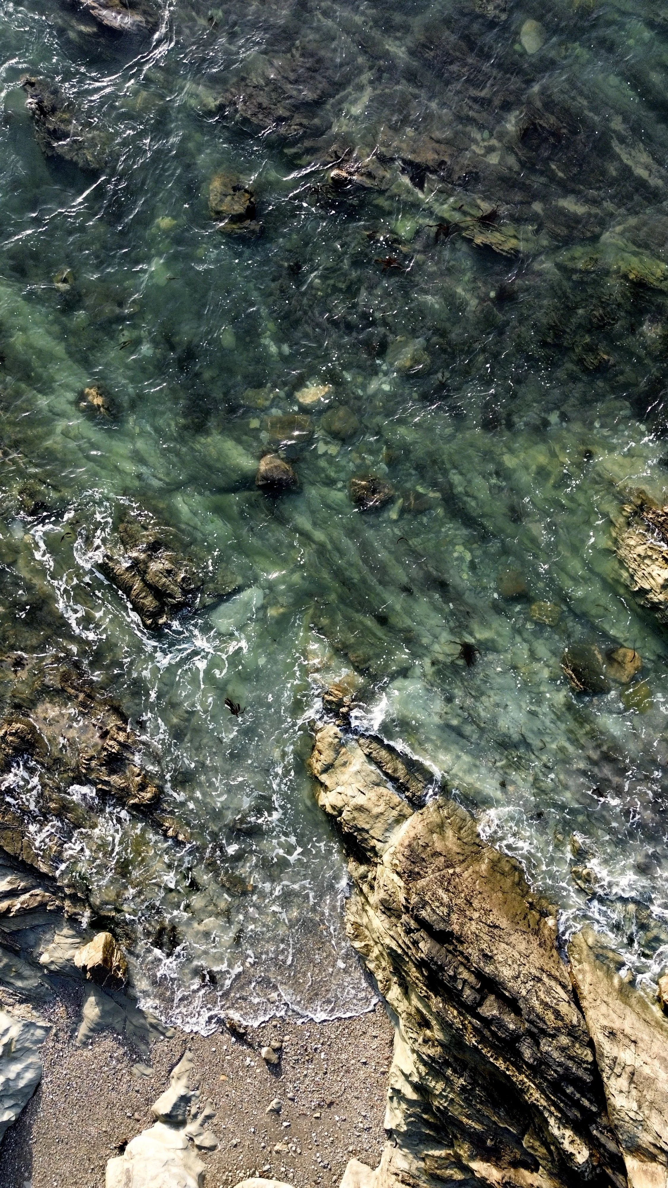 Clear ocean water with visible rocks and submerged cliffs near the shoreline, with small waves crashing against the rocks.