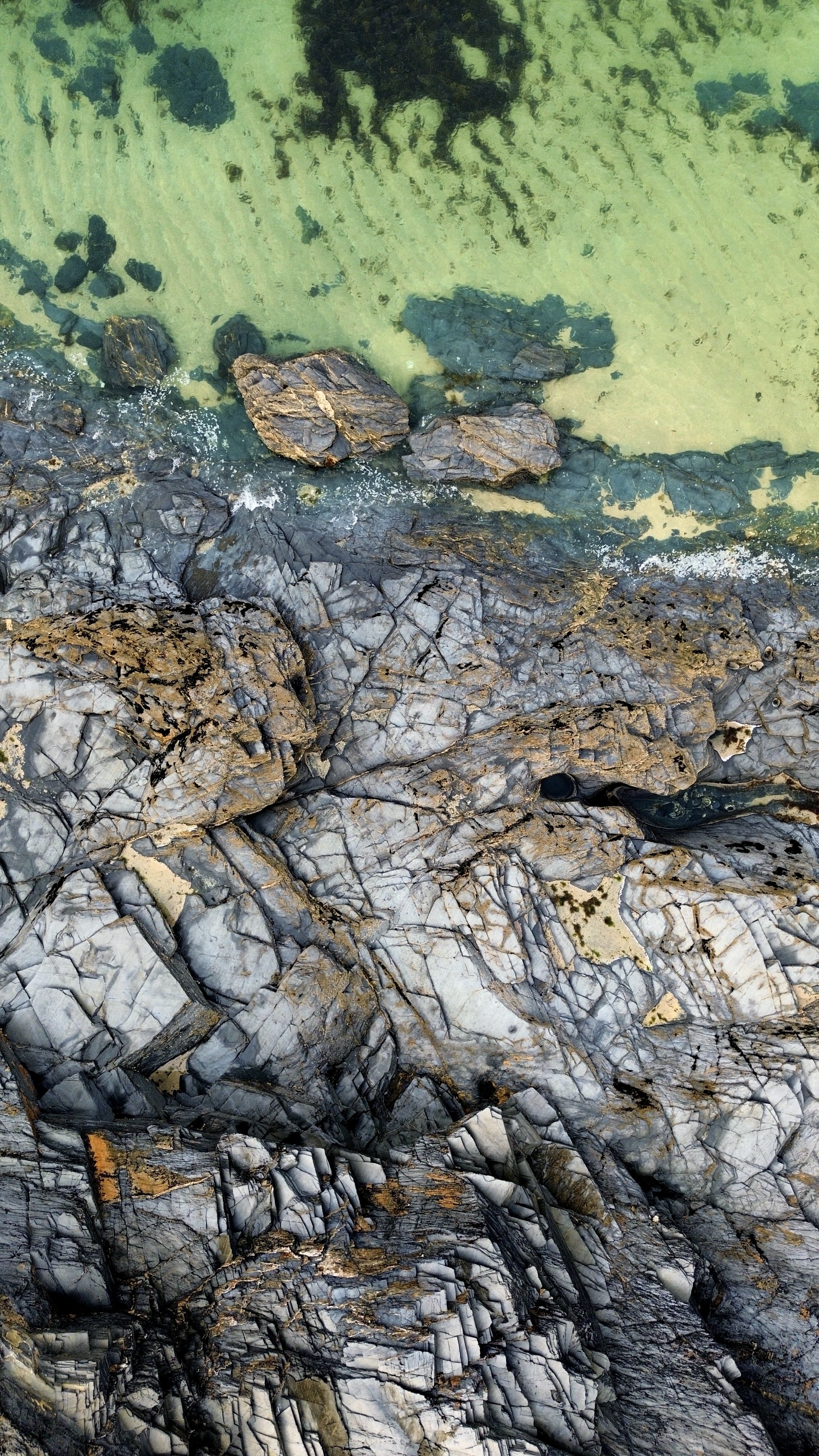 Aerial view of a rocky shoreline with large boulders, green water, and underwater rocks visible beneath the surface.