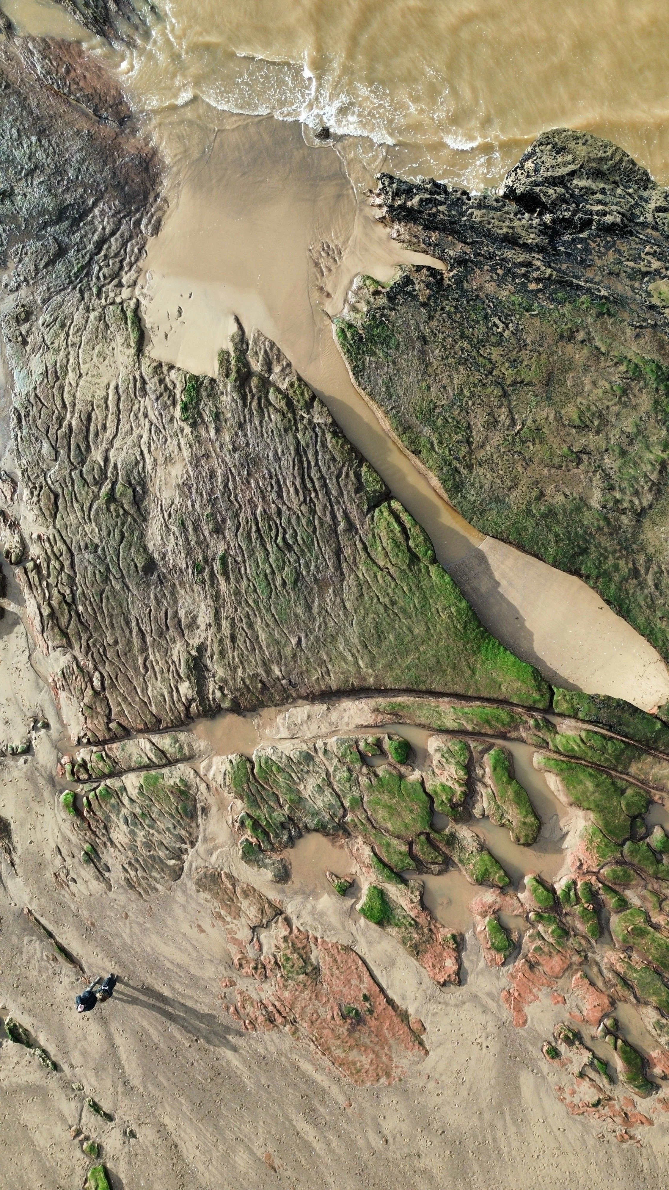 Aerial view of a rocky coastline with sandy beaches, green moss-covered rocks, and the ocean waves crashing against the shoreline.