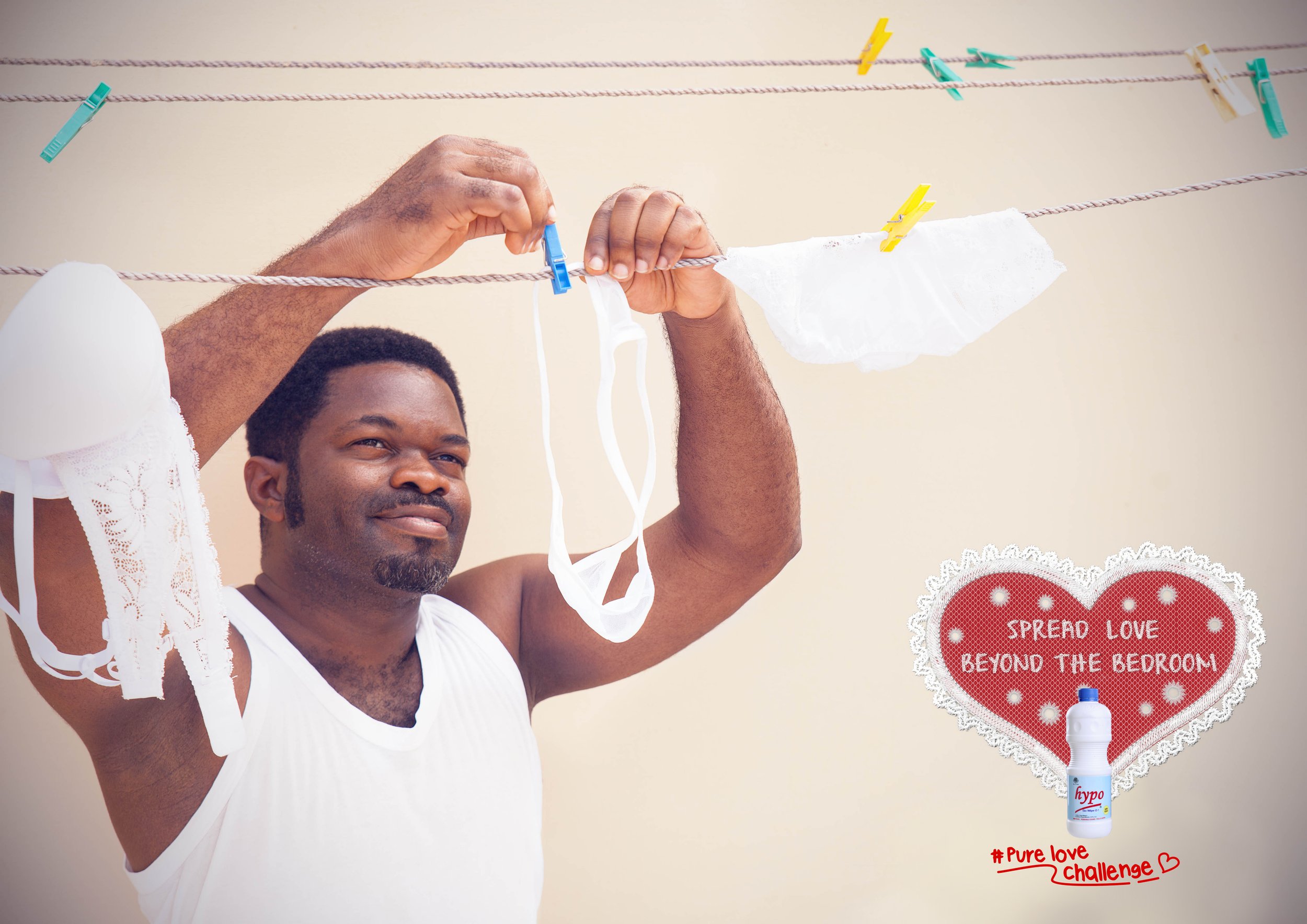 A man hanging laundry on a clothesline with clothespins. There is a decorative heart-shaped sign in the background with the message 'Spread Love Beyond the Bedroom.' A bottle of hypoallergenic laundry detergent labeled 'hypo' is placed below the sign, and the hashtag '#PureLoveChallenge' with a heart symbol is written in red at the bottom right corner of the image.