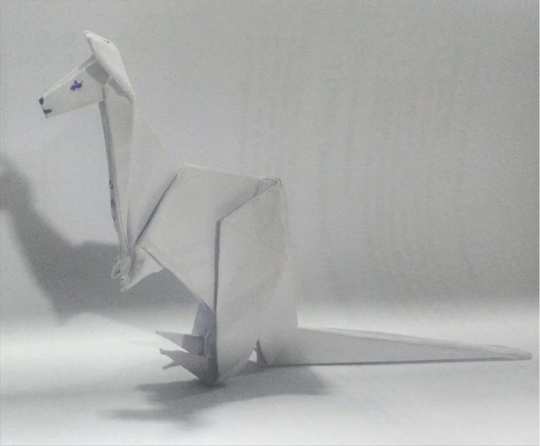 A paper origami creation resembling a dragon or dinosaur, placed on a white surface against a plain white wall. Sodiq Sheu Olashile, Best Art Director in Nigeria.