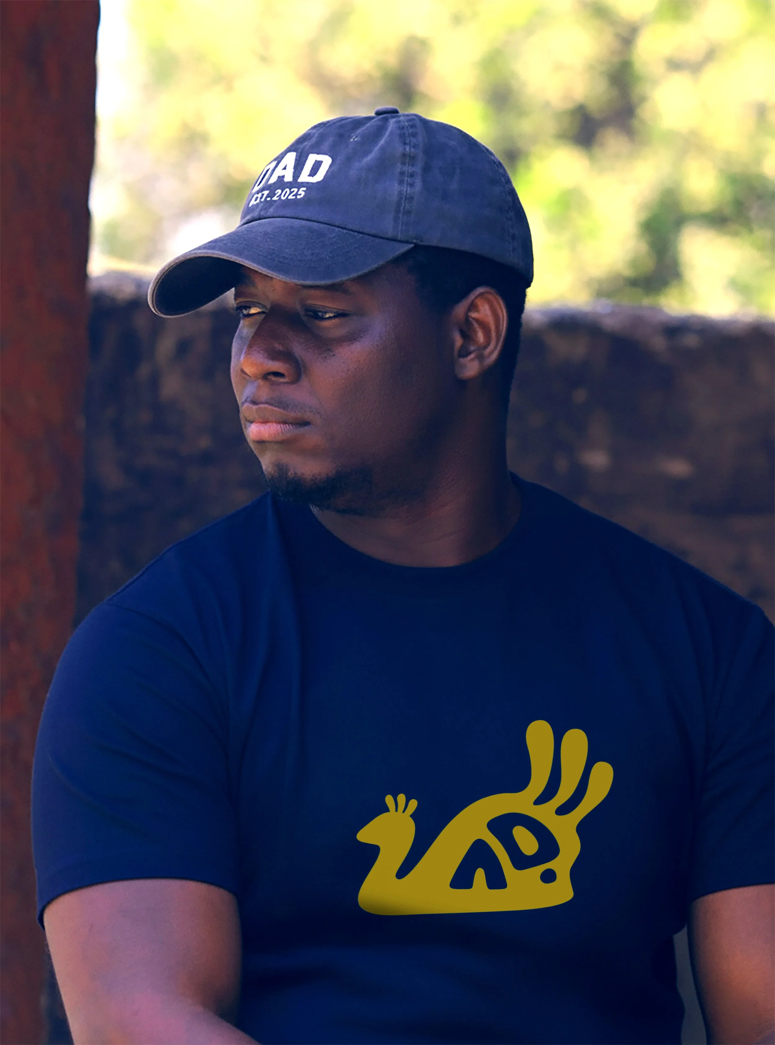 A young Yoruba man wearing a dark blue baseball cap with white text and a black T-shirt with a yellow Ayinde Okin logo design on it, standing outdoors with blurred greenery in the background. Yoruba Artist.
