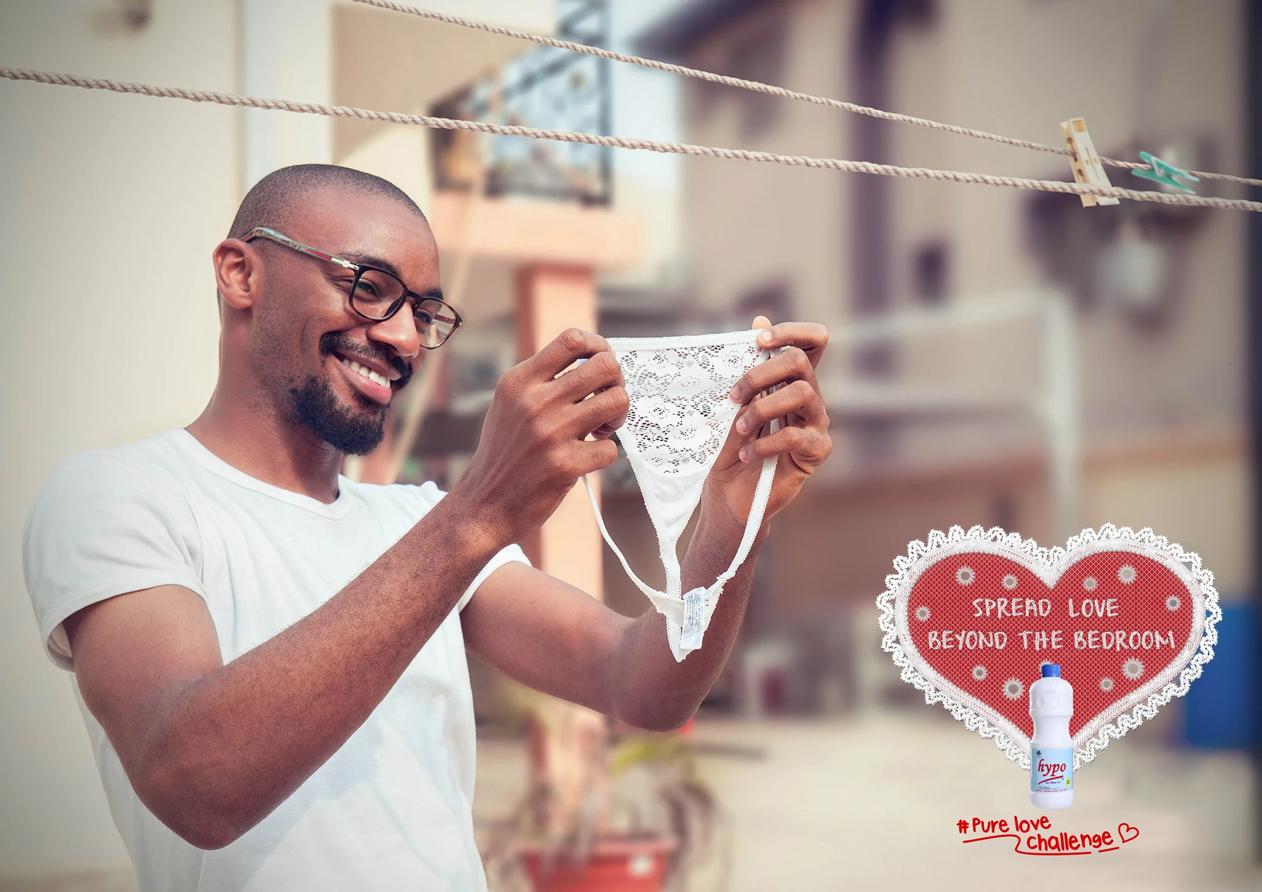 A man with glasses and a beard smiling while holding a lace thong. There is a red heart-shaped sign with white lace trim that reads "Spread Love Beyond the Bedroom" and a small bottle of hand sanitizer attached to it, with the hashtag "Pure Love Challenge" written below.