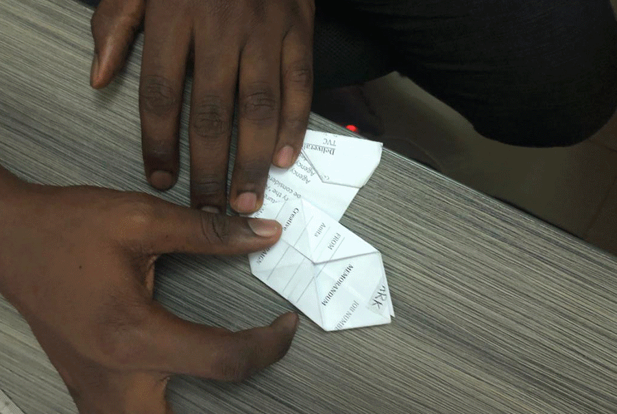 Two people folding a piece of paper into an origami shape on a wooden table. Sodiq Sheu Olashile, Best Art Director in Nigeria.